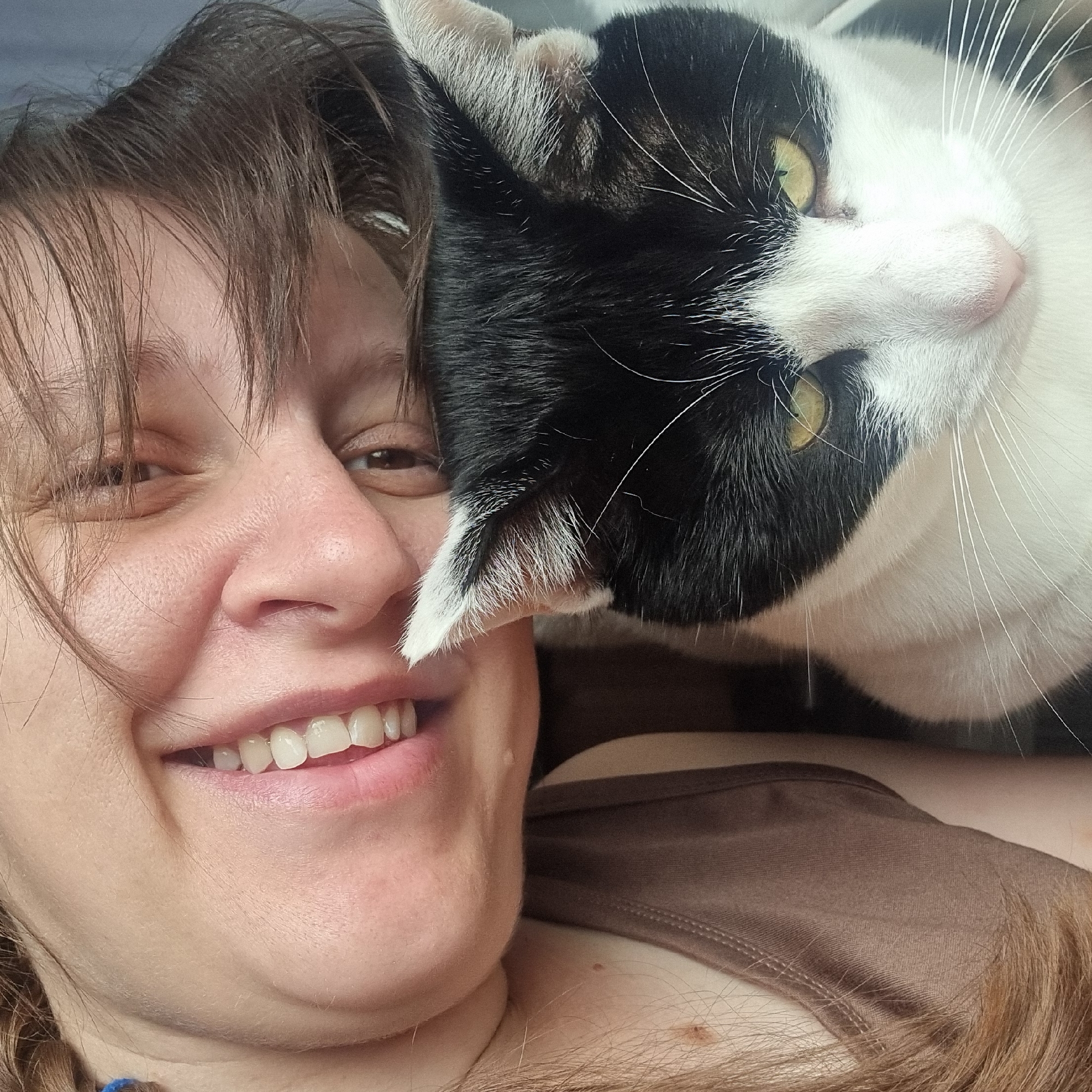 The image shows a close-up, overhead shot of a person and a cat. The person, who appears to be smiling, is positioned on the left side of the frame, with their face tilted towards the camera. Their hair is brown and appears somewhat messy. The cat, a black and white feline, is in the lower right portion of the image, looking directly at the camera with yellow eyes. The cat's fur is visible in detail, particularly around its face and ears. The person is wearing a brown tank top. The background is a plain, dark color. The image was taken in Dunnae-myeon, South Korea.