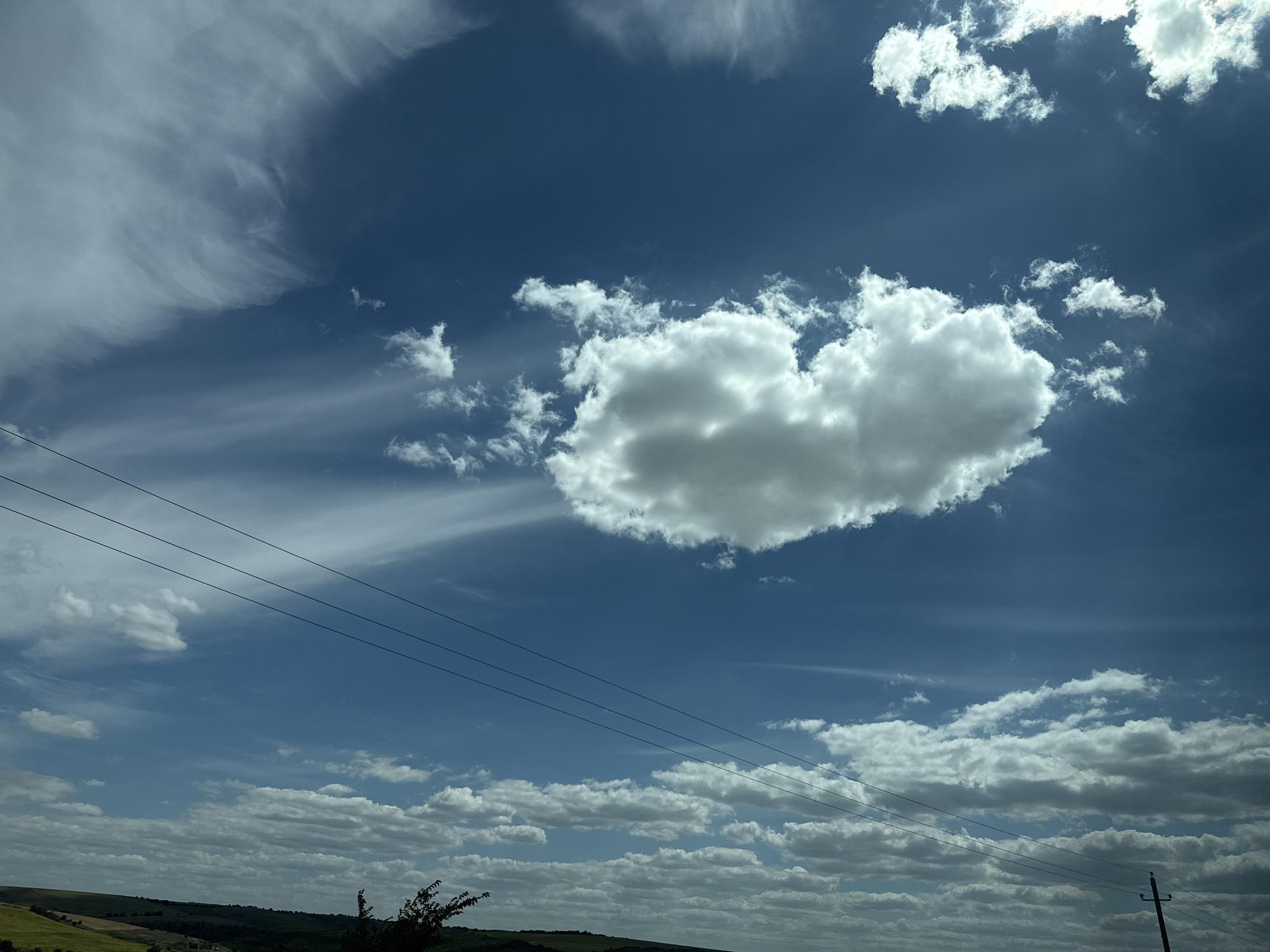 The image captures a wide-angle view of a bright, partly cloudy sky above a rural landscape. Three parallel power lines stretch horizontally across the lower portion of the frame, cutting through the expanse of blue sky dotted with various cloud formations. The clouds range from wispy cirrus clouds that streak across the upper left to larger, more voluminous cumulus clouds clustered in the lower half and towards the right.
Below the sky, the landscape is characterized by rolling hills with patches of green and yellow fields, suggesting agricultural land. A few scattered trees and a dark silhouette of vegetation are visible near the bottom edge, with a single utility pole on the far right. The lighting indicates it is daytime, with the sun casting shadows and illuminating the clouds from above. The overall impression is one of an open, natural environment under a dynamic sky.
The image is stated to be located in Grădişte, Moldova.