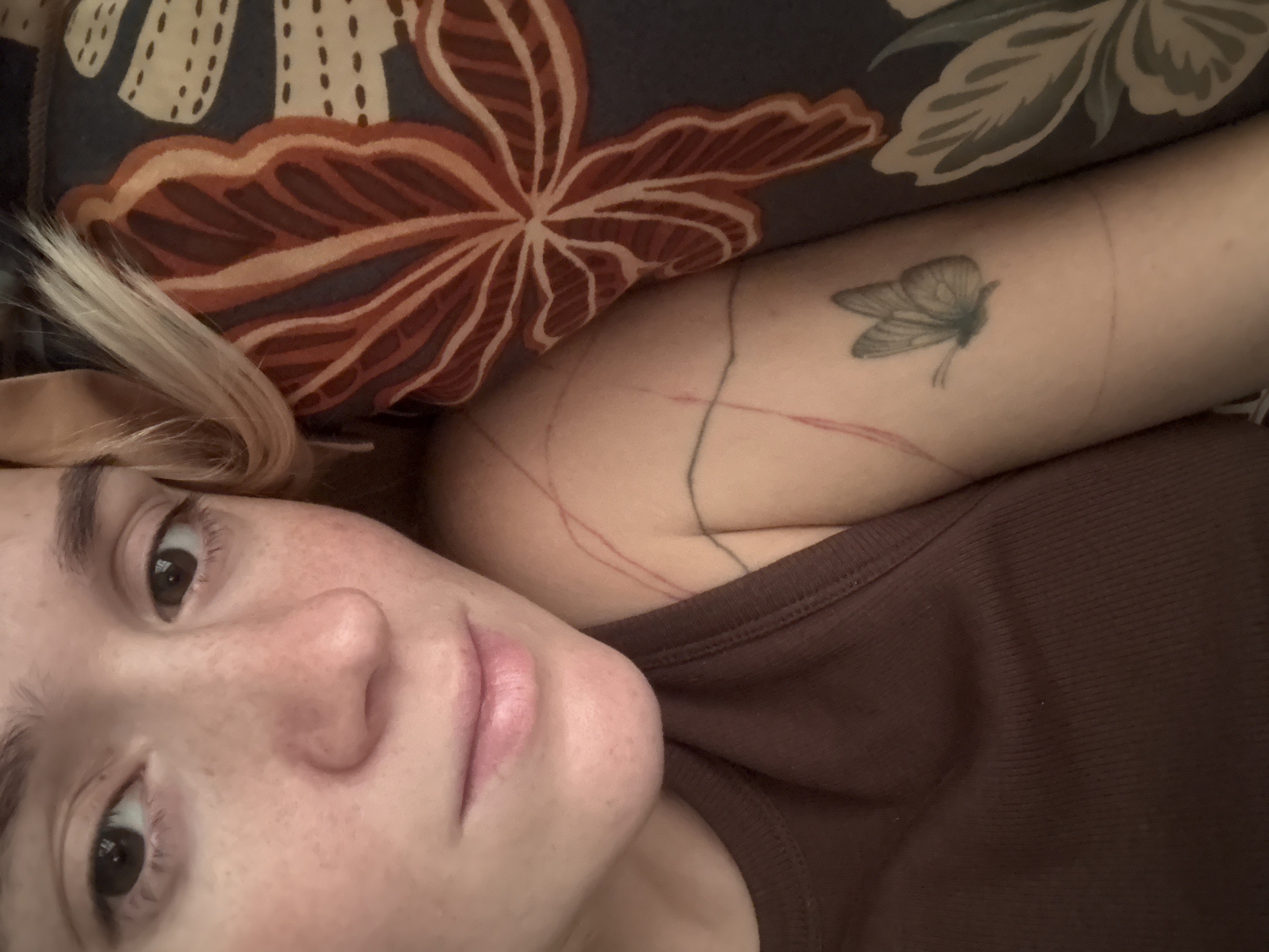 A close-up, overhead shot shows a person with fair skin and freckles lying on a bed. The person's face is partially visible, with their eyes, nose, and mouth in focus. Their hair is blonde and light-colored, and it drapes over the right side of their head. The person is wearing a dark brown tank top. On their left arm, there is a small butterfly tattoo and some red markings. Behind the person, a pillow with a floral pattern is visible. The background appears to be a bedroom setting. There is no information in the image that indicates the location in the city of Nizhny Novgorod, Russia.