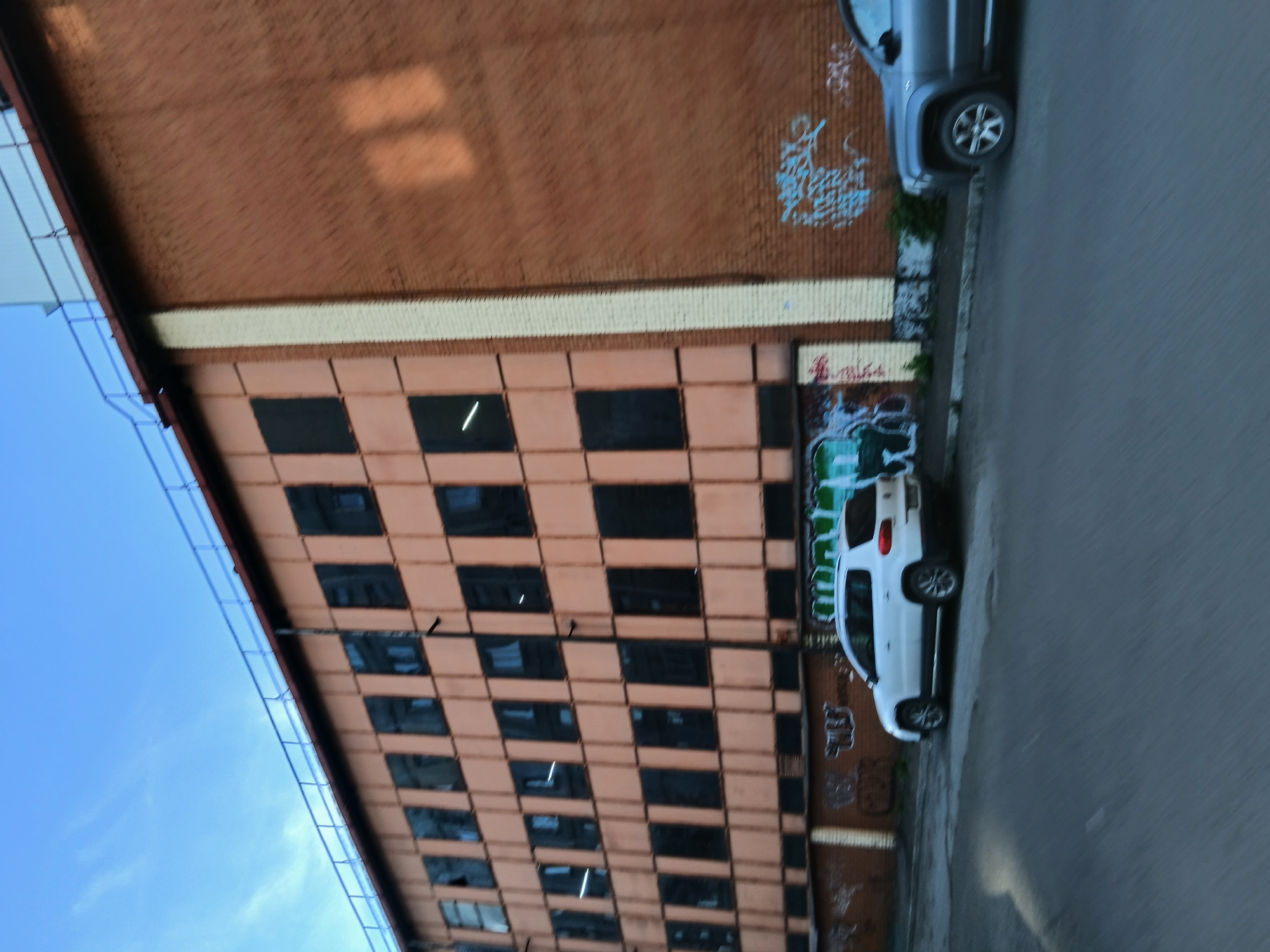 A motion-blurred outdoor, eye-level, medium shot captures an abandoned, multi-story brick building with a light orange facade. The building has rows of dark, boarded-up windows. The roofline features a blue railing. To the right, a section of the building is made of red brick, adorned with blue and white graffiti. In front of the building, parked on the asphalt, are a white SUV on the left and a dark SUV on the right. The sky above is blue with wispy clouds. The foreground is a blurred asphalt road.