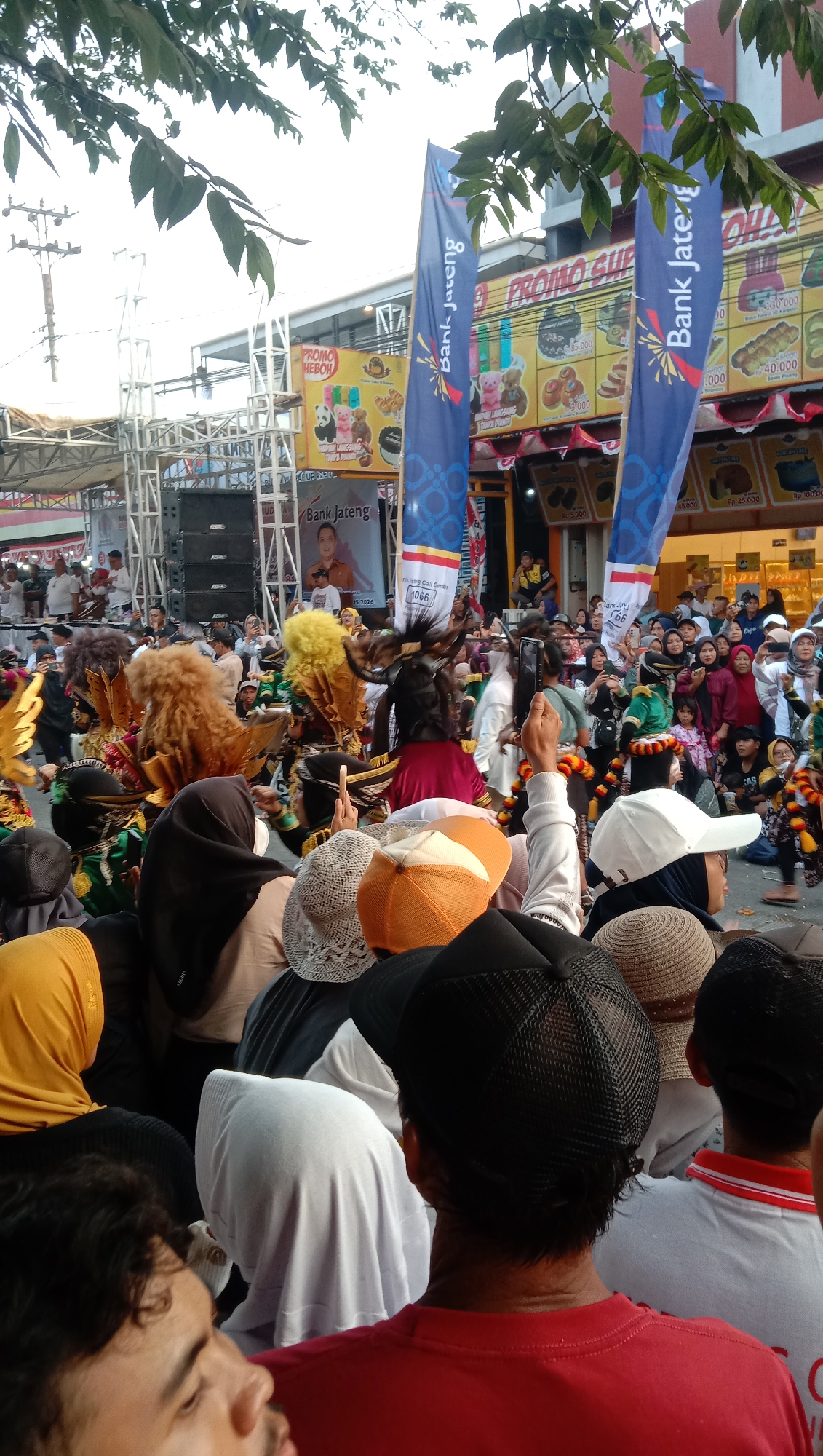 The image depicts a lively outdoor event, possibly a festival or parade, in the city of Jatisawit, Indonesia. A crowd of onlookers, many wearing headscarves and caps, are gathered in the foreground, facing a central performance area. In the middle ground, performers in elaborate, colorful costumes, some with large, feathery headdresses and what appear to be animalistic masks, are engaged in a procession. Behind them, a stage or platform with large speakers is visible, suggesting a musical or theatrical performance. Two large blue banners with text that appears to be "Bank Jateng" are prominently displayed, indicating a sponsor. The background shows shops and buildings, some adorned with promotional banners, adding to the festive atmosphere. The presence of trees and the overall lighting suggest it is daytime.
