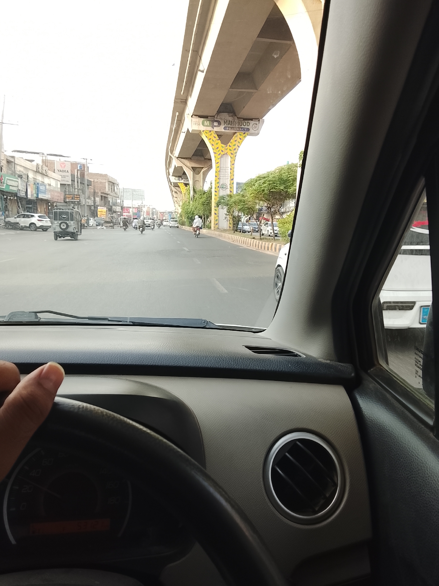 The image shows a view from inside a car driving on a road in Gulgasht Colony, Multan, Pakistan. The driver's hand is visible on the steering wheel. The road is relatively straight with an elevated concrete structure, possibly a metro line, running above it. Several vehicles, including cars and motorcycles, are visible on the road ahead. Buildings and shops line the side of the road. The weather appears bright and slightly hazy.