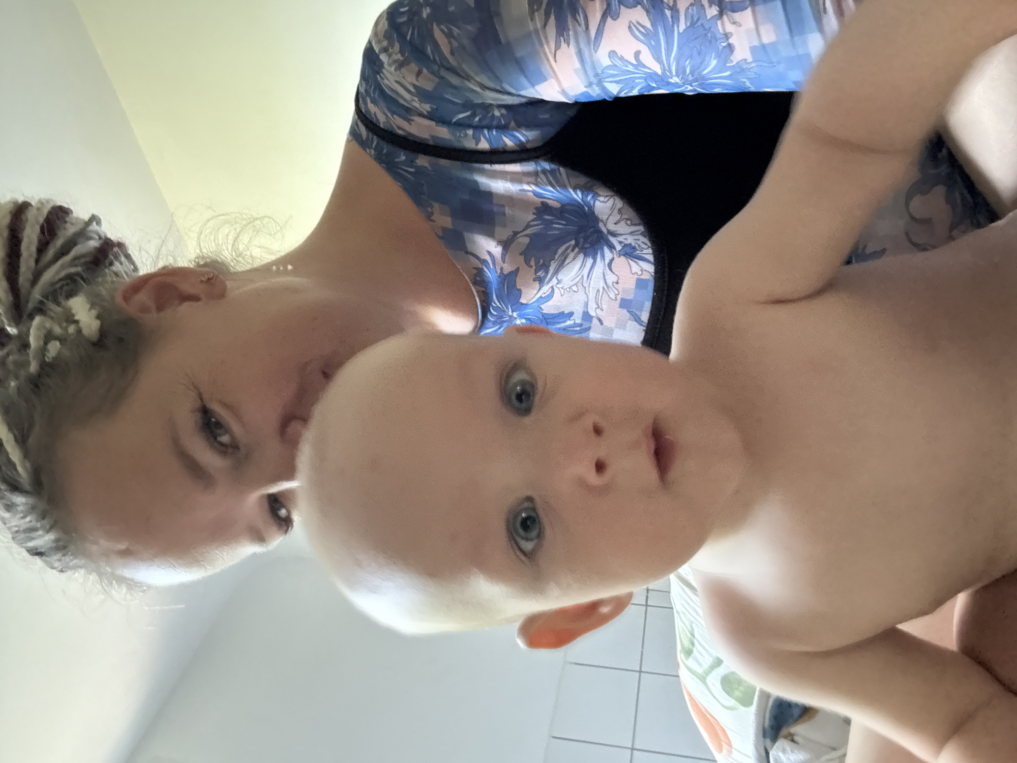 In Sochi, Russia, a woman with braided hair is taking a selfie with a bald baby. The baby is in the foreground, looking at the camera with wide blue eyes and an open mouth. The woman is behind the baby, her face partially obscured as she leans in. She is wearing a blue and white floral print top over a black camisole. The background shows a tiled wall and a pillow. The lighting suggests an indoor setting.