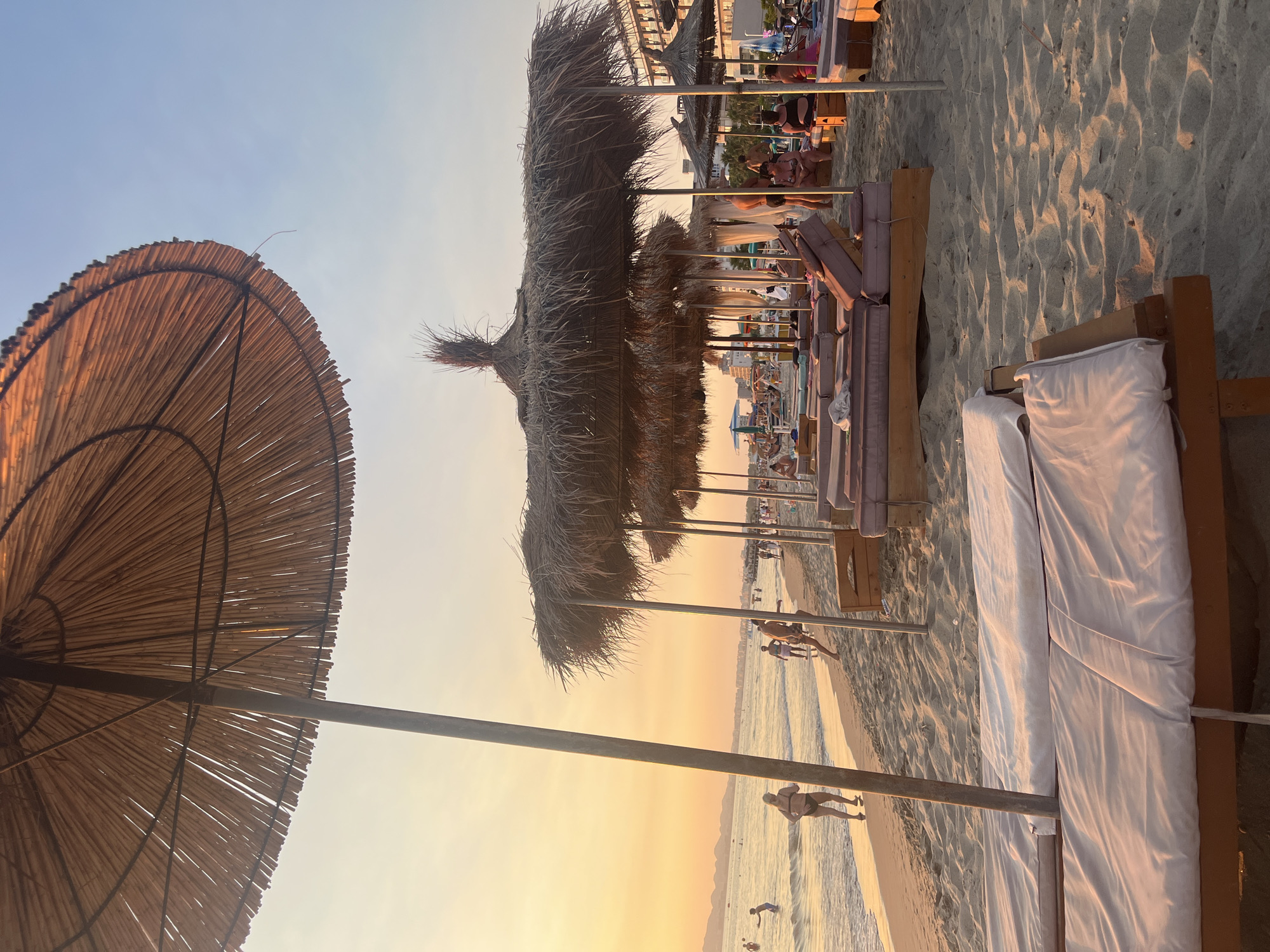 The image depicts a beach scene at sunset in Bashkia Durrës, Albania. The foreground is occupied by several empty lounge chairs with white covers, positioned on the sandy beach. Behind these chairs, a row of thatched umbrellas provides shade over more lounge chairs, some of which are occupied by people. In the middle ground, the gentle waves of the sea lap against the shore, and several individuals are seen walking or wading in the water. The background shows a distant coastline with buildings and a hazy horizon. The sky is a warm gradient of orange and blue, indicative of a sunset. The overall atmosphere is one of a tranquil beach day coming to a close.