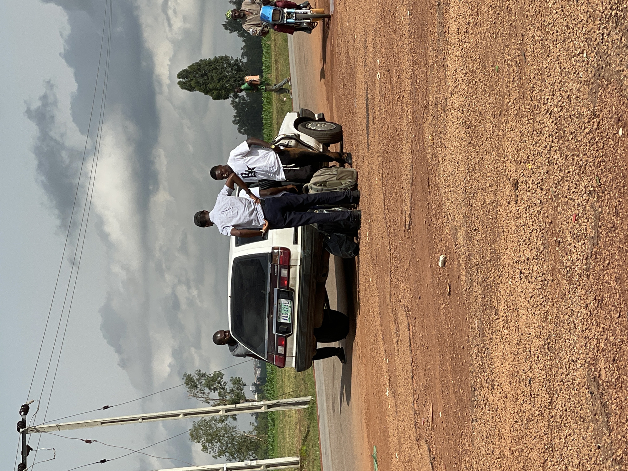 In Wumat, Nigeria, on a cloudy day, a white sedan is parked on a dirt road next to a paved road. Three men are standing near the car, with luggage visible around them. One man, wearing a grey shirt and dark pants, is looking towards the back of the car. Another man in a white shirt and dark pants is looking away from the camera. The third man, wearing a white t-shirt with a black graphic and dark pants, is facing the camera. In the background, a person walks by a field, and a man rides a motorcycle. Power lines and poles are present on the left side of the image.