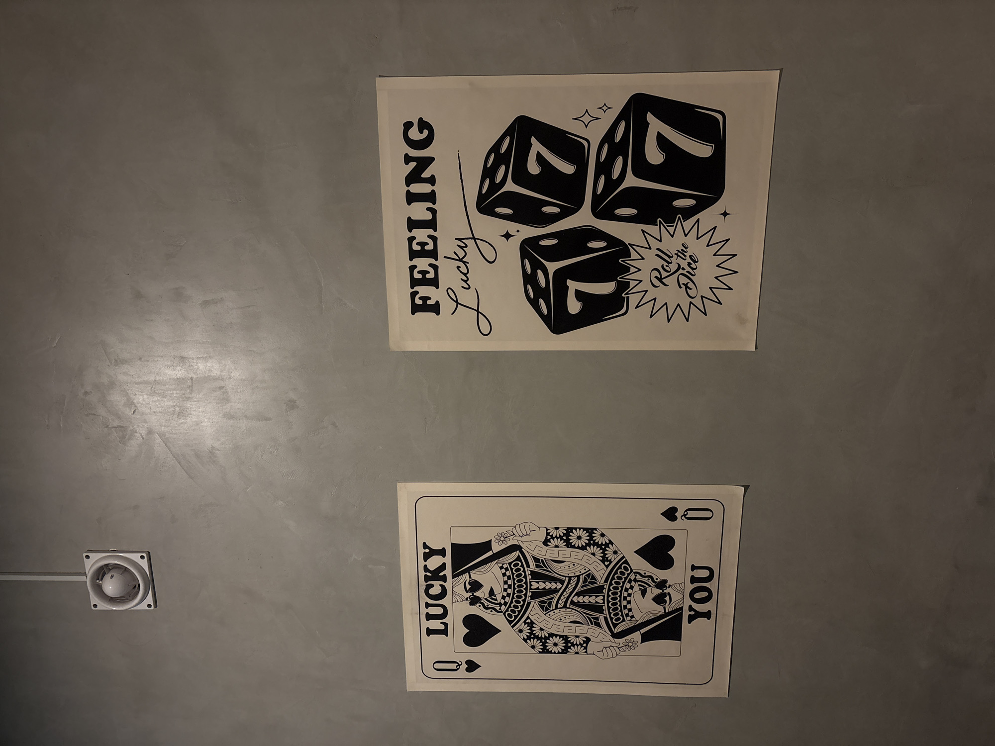 The image shows two posters on a gray wall. The top poster has the words "FEELING Lucky" and an illustration of three dice, each showing the number 7. The bottom poster is a Queen of Hearts playing card with the words "LUCKY" and "YOU" on either side. A ceiling fan is visible in the bottom left corner of the image. The exact location in San Andrés Cholula, Mexico, cannot be determined from this image.
