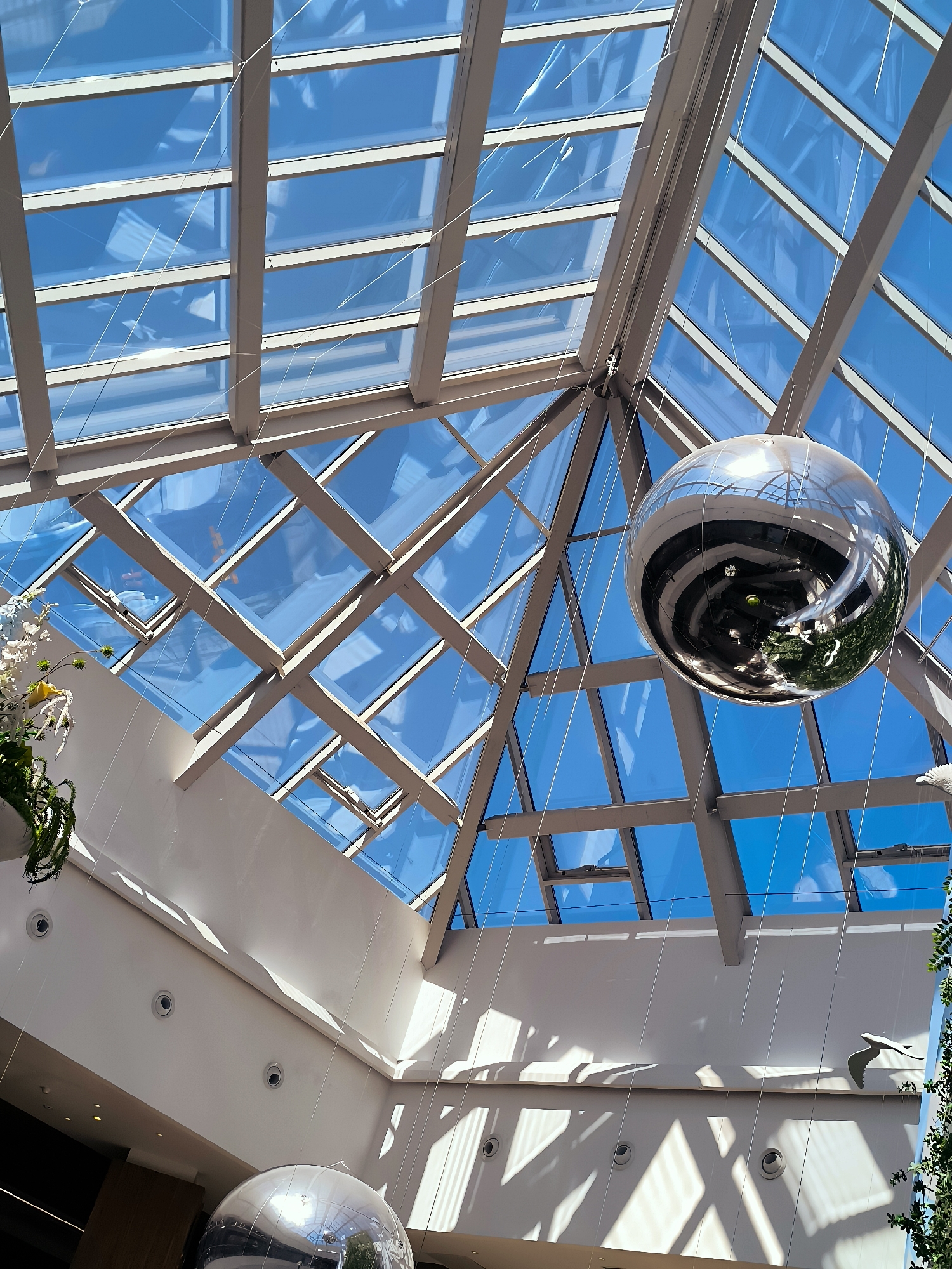 The image captures a view looking upwards towards a large, modern glass atrium ceiling. The ceiling is constructed with a grid of metal beams and glass panels, allowing bright blue sky and sunlight to filter through. Suspended within the atrium are several large, spherical, reflective decorations. One prominently features a spiral pattern. The walls of the building are painted a light color and are illuminated by sunlight creating geometric shadow patterns. There are circular light fixtures visible on the walls. A potted plant with green foliage is visible on the left side of the frame, and a white bird-shaped sculpture is seen on the right. The setting appears to be an indoor public space, possibly a shopping mall or a government building in Qarağandı city administration, Kazakhstan.