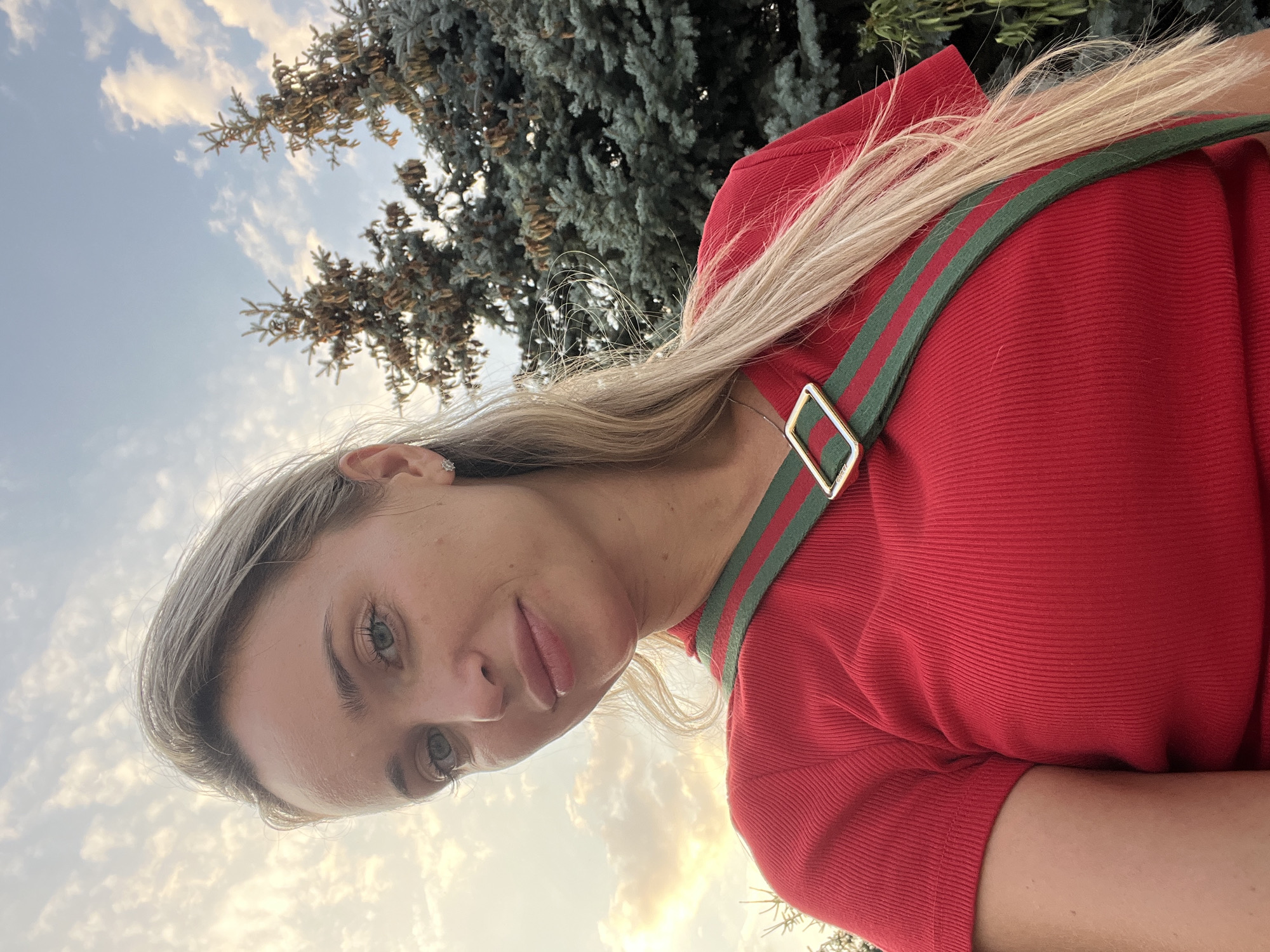 A blonde woman stands outdoors, facing the camera. She is wearing a red, ribbed, short-sleeved shirt with a green and red striped strap diagonally across her chest. The strap has a gold rectangular buckle. Her blonde hair is worn down and falls past her shoulders. She has light-colored eyes and is looking directly at the camera. Behind her, there are two evergreen trees and a sky with scattered white clouds. The location is identified as Fontanka, Ukraine.