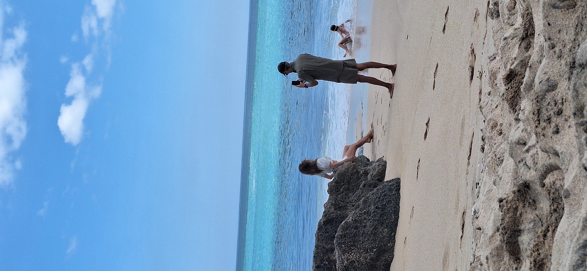 In Kuta Selatan, Indonesia, on a sandy beach, a man wearing a dark cap and olive-green short-sleeved shirt and shorts stands on the shore, holding a black smartphone up to take a picture. To his left, a woman with long, light-brown hair sits on a large, dark gray rock, wearing a white top and white shorts. She is looking away from the camera. Further in the background, a person in the shallow turquoise water is lying on their side, appearing to be taking a photo or video with a smartphone. The ocean stretches to the horizon under a clear blue sky with scattered white clouds. The beach is composed of light-colored sand with footprints visible.