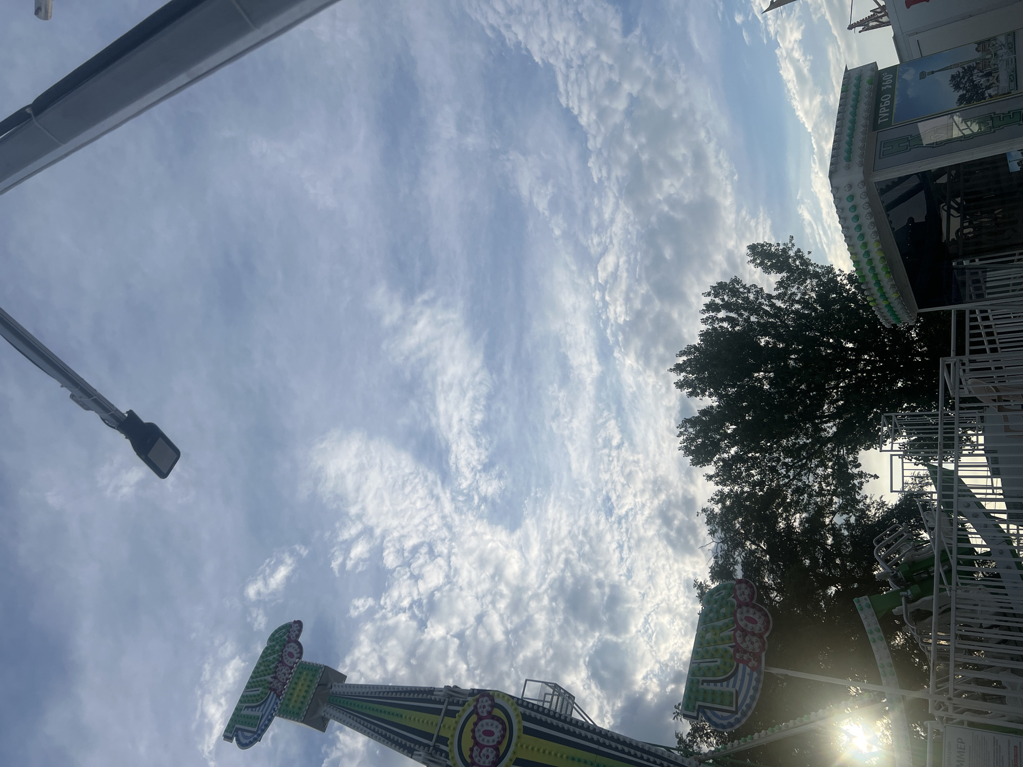The image captures a fairground ride in Peresyp, Russia, under a partly cloudy sky. The ride is a tall, green and yellow structure with "TURBO 360" displayed prominently. In the foreground, part of the ride's platform and fencing is visible, along with trees. The sun is shining through the clouds, creating lens flare on the left side of the image. The scene appears to be set up for a recreational event.