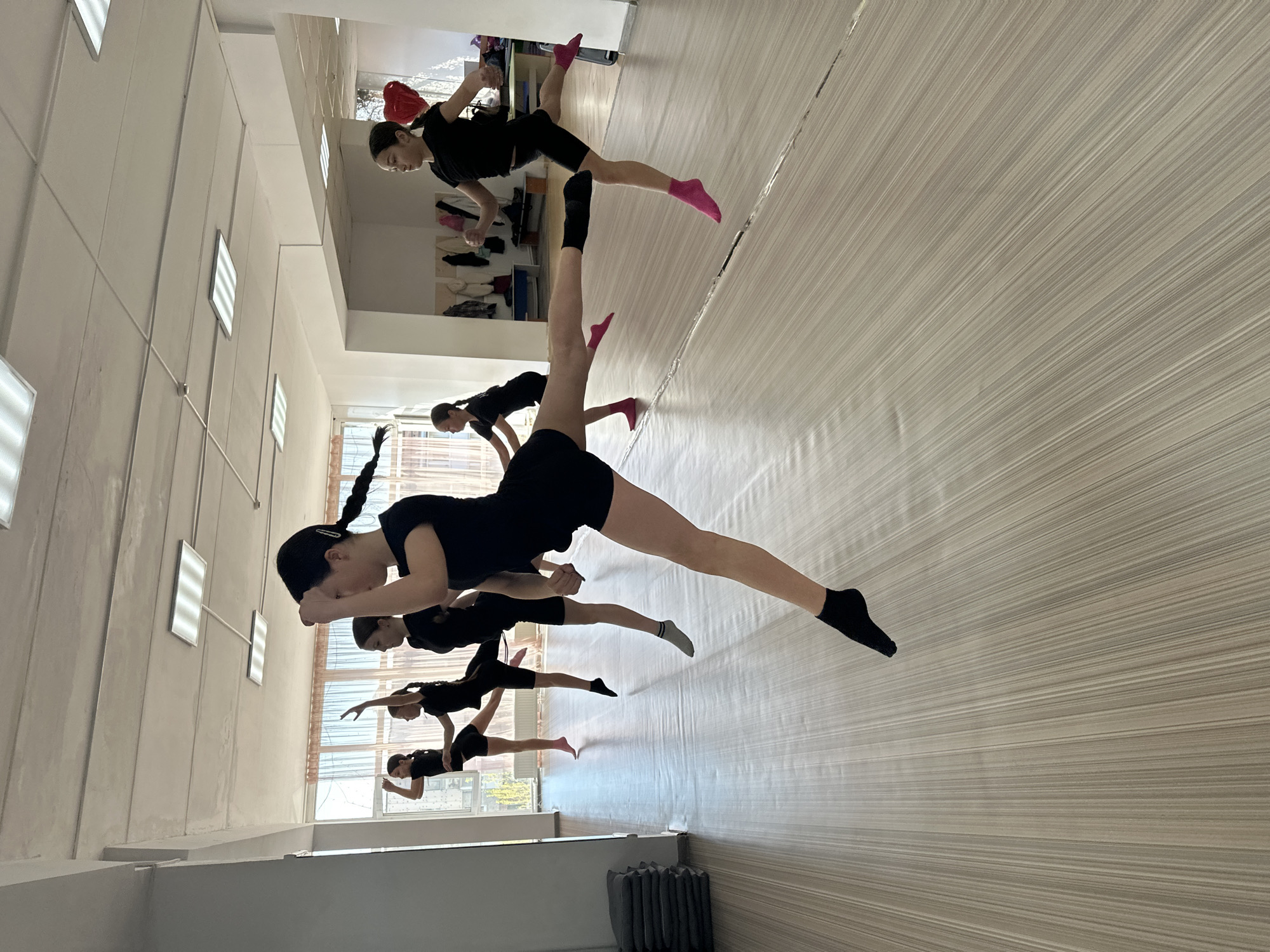 A group of young female dancers wearing black leotards, shorts, and socks (mostly pink or black) are practicing a dance routine in a brightly lit studio. The dancers are suspended in mid-air, performing split leaps, with one leg extended forward and the other back. The studio has a light-colored, horizontally striped floor, white walls with rectangular light fixtures, and a large mirror along one side, reflecting the dancers and the room. In the background, visible through a window, is an indistinct urban scene, possibly in Votkinsk, Russia.