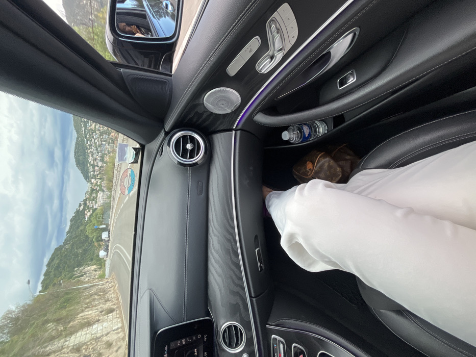 The image is taken from the interior of a car, likely a Mercedes Benz, driven by a person wearing white pants. The car is parked and the driver is looking ahead through the windshield. The image is taken from the passenger seat, with the driver's leg visible on the passenger side. There are several objects in the vehicle: a water bottle in a cup holder, a purse on the floor, and a control panel for the seat and windows. The driver has the windows rolled down and there is a view of the road and surrounding environment through the windshield. The road is winding and there are trees and hills in the background. The location is the coastal city of Villefranche-sur-Mer, France, based on the coordinates. The image is captured during the day with overcast skies.