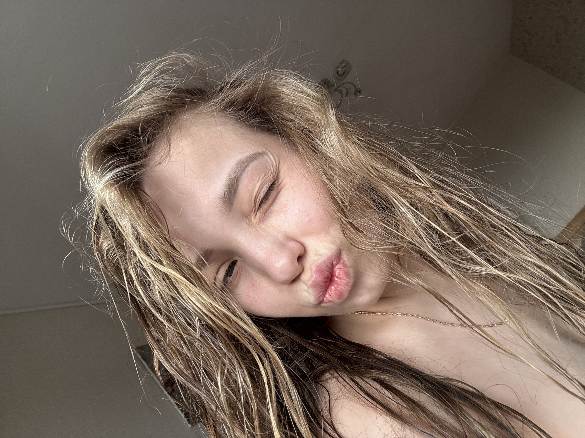 A close-up, angled shot shows a young woman with long, damp, blonde and brown hair puckering her lips in a kissing gesture. She has one eye closed in a wink. She wears a thin, gold chain necklace. The background appears to be an interior room with a plain, off-white wall and a textured ceiling visible in the upper right corner. The location, Chaikovsky, Russia, is not evident in the image.