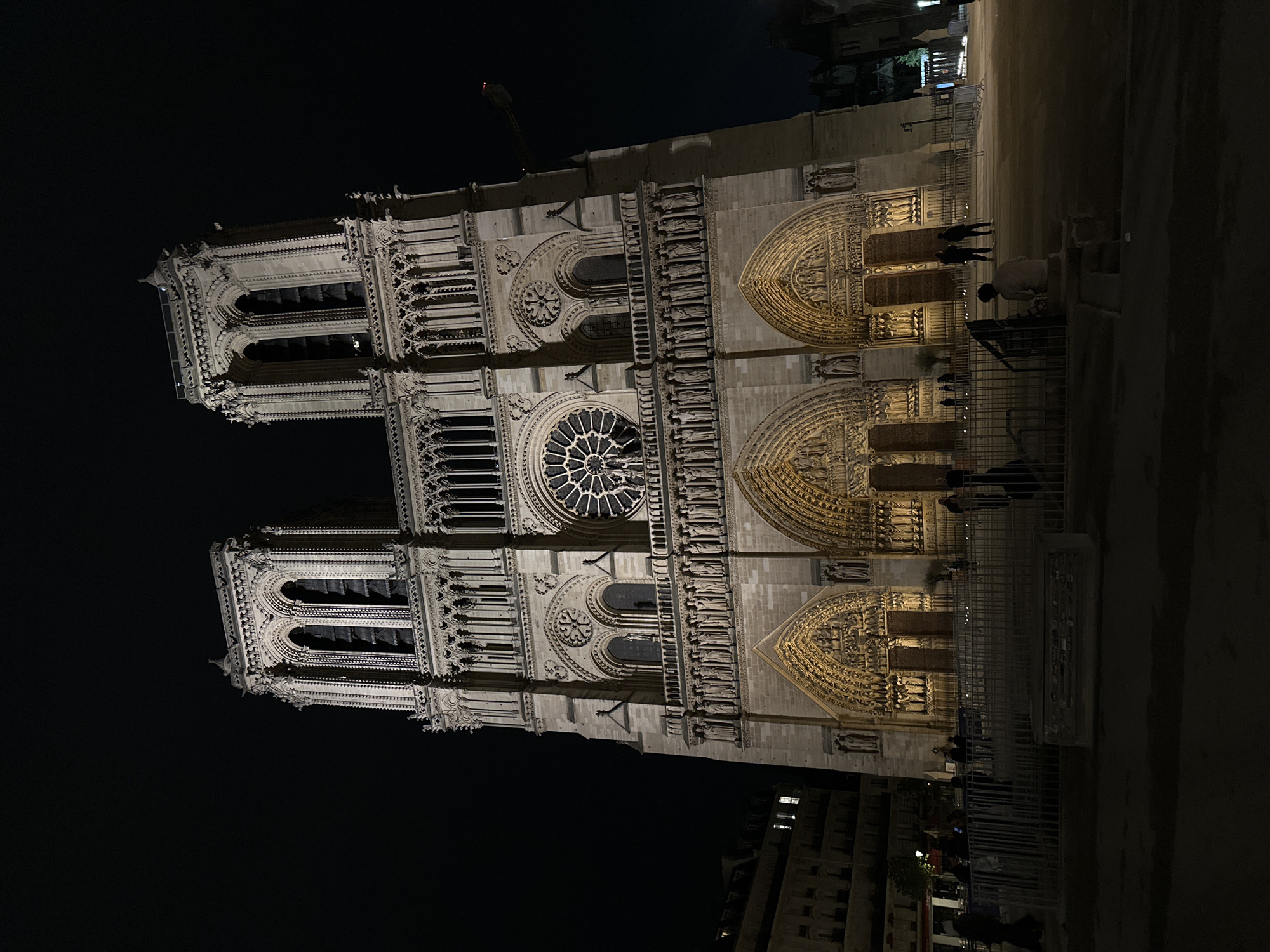 The image shows the facade of Notre-Dame Cathedral in Paris, France, at night. The cathedral's two towers and central rose window are visible, illuminated by artificial light. A construction crane is silhouetted against the dark sky above the right tower. A metal fence separates the cathedral from a public area where people are standing and walking. Buildings are visible behind the cathedral.