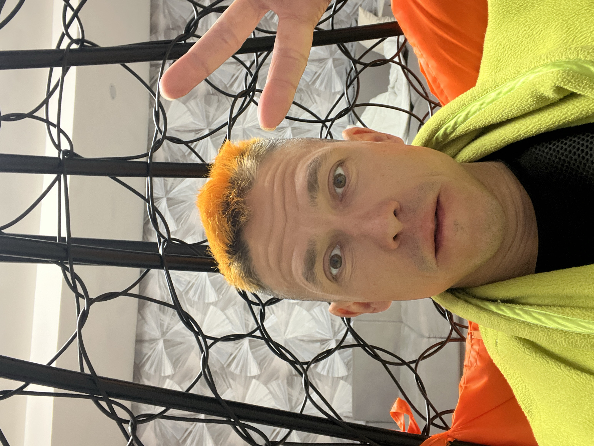 A close-up, tilted selfie features a fair-skinned person with short, bright orange hair. They are wearing a black shirt and a bright green fleece jacket layered over a bright orange article of clothing. The person is making a peace sign with their right hand. In the background is a black metal grid with a patterned white wall visible behind it. The exact location in the city of Chaikovsky, Russia is not discernible from the provided image.