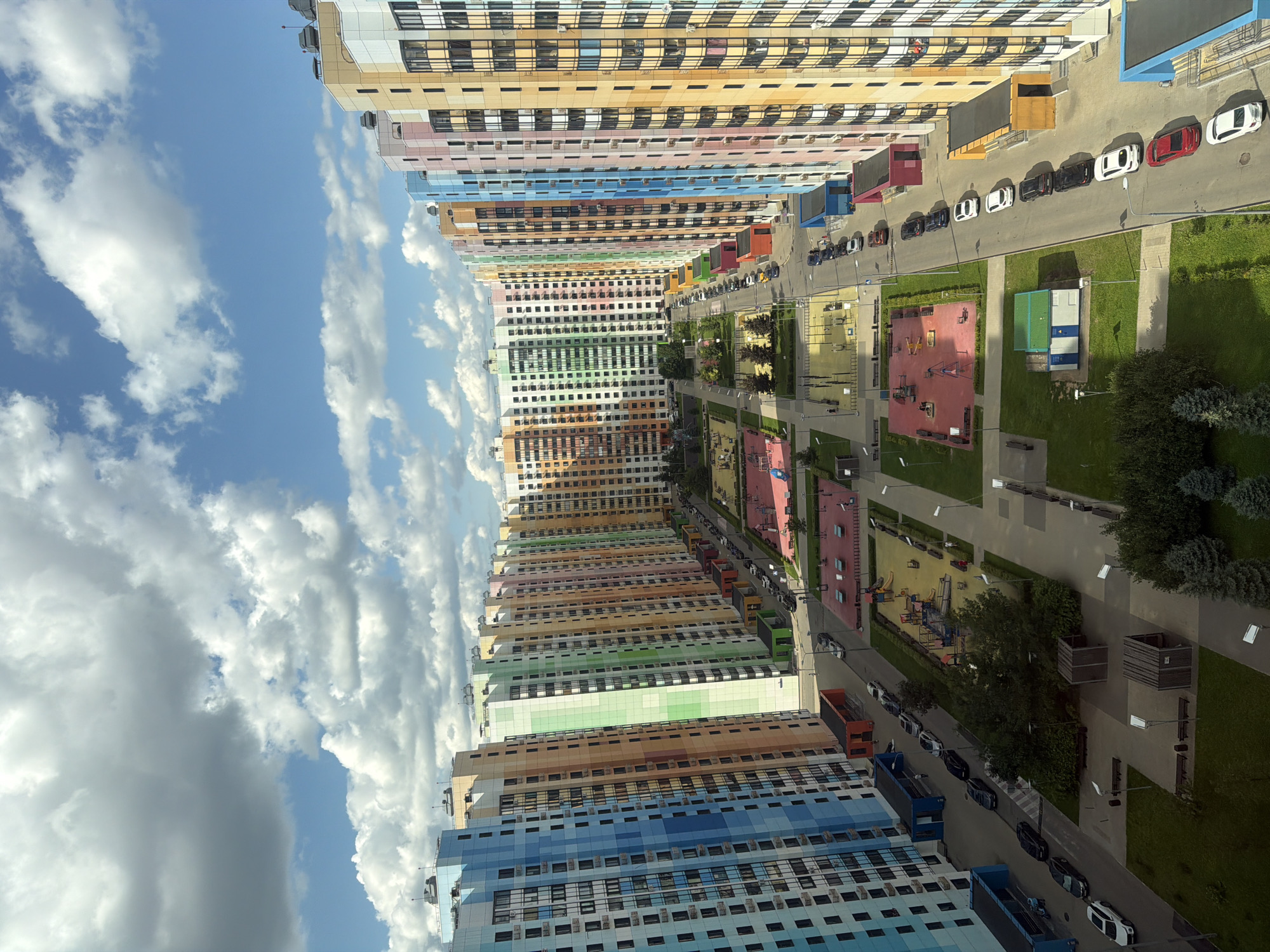 The aerial image depicts a densely populated residential area with rows of towering, multi-colored apartment buildings. The buildings are arranged in a courtyard-like fashion, with a central area containing playgrounds and recreational spaces. A road runs between the apartment blocks, lined with parked cars. The sky is a vibrant blue, dotted with fluffy white clouds, indicating fair weather. The overall impression is one of a modern, planned community with a focus on family-friendly amenities. The colors of the buildings are predominantly bright and varied, including blue, green, pink, orange, and yellow, which creates a cheerful and lively atmosphere. There are no visible individuals in the frame, and the scene appears to be devoid of any unusual or hazardous activity.