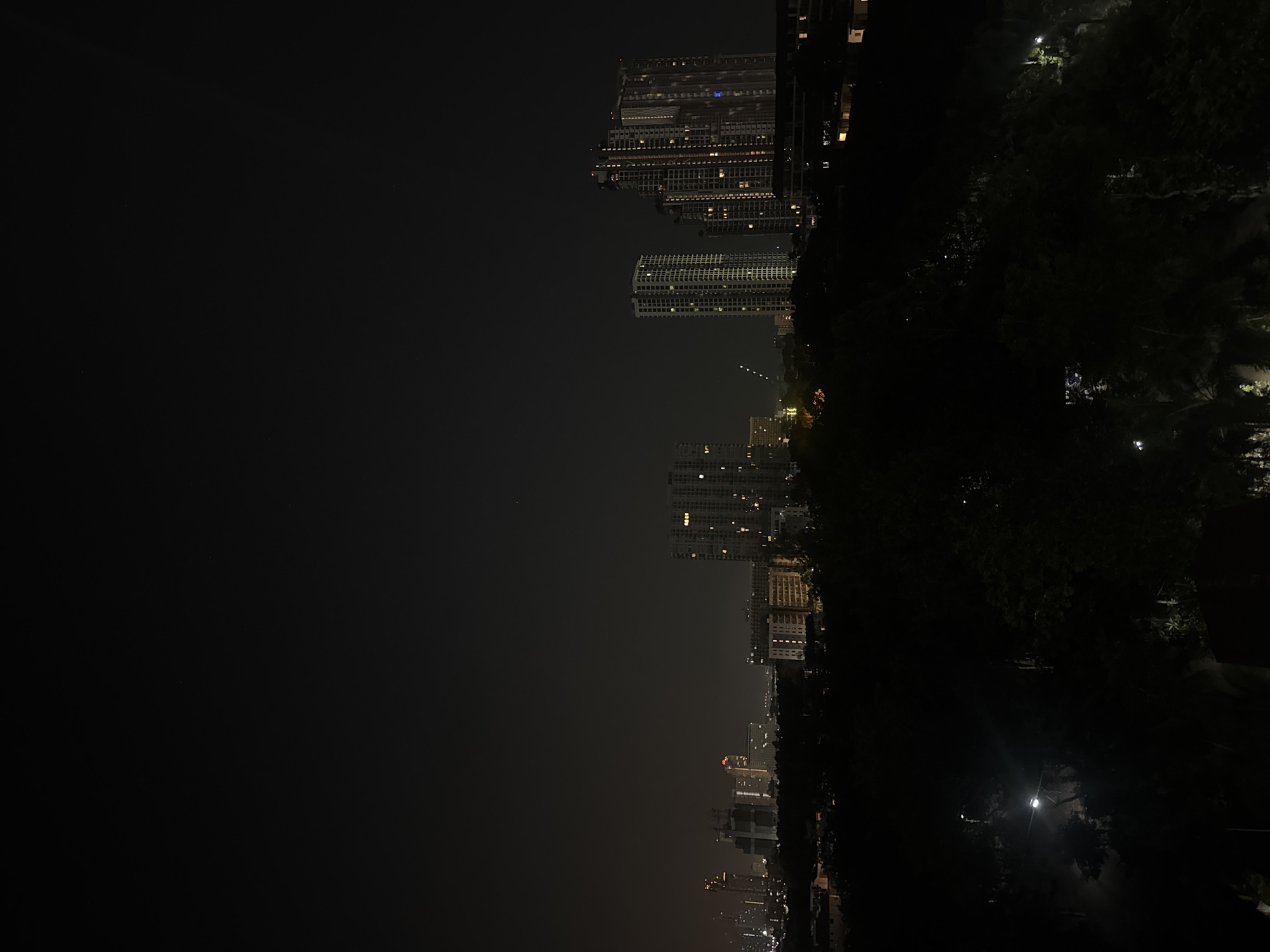 The image captures a panoramic night view of a city skyline, likely Pattaya City, Thailand, as indicated by the context. The dominant feature is a vast, dark sky, occupying a significant portion of the left side of the frame, suggesting a late hour. The sky transitions from deep black at the top left to a slightly lighter, hazy dark gray towards the horizon where urban lights begin to appear.
On the right side, a dense collection of high-rise buildings stands tall, their dark silhouettes punctuated by numerous illuminated windows. These lights, appearing as small, bright squares and rectangles, indicate the presence of residential or commercial activity within the multi-story structures. Several distinct clusters of buildings are visible, with varying heights and architectural patterns.
In the immediate foreground, along the bottom edge and extending slightly upwards on the far right, is a mass of dark, dense foliage, likely trees, suggesting a natural buffer zone such as a park or green area. A few faint streetlights or other light sources are scattered within this dark greenery, casting subtle glows. The overall atmosphere is serene and urban, with a hint of atmospheric haze obscuring the distant details of the city. No specific activities or events are discernible, and there is no visible text.