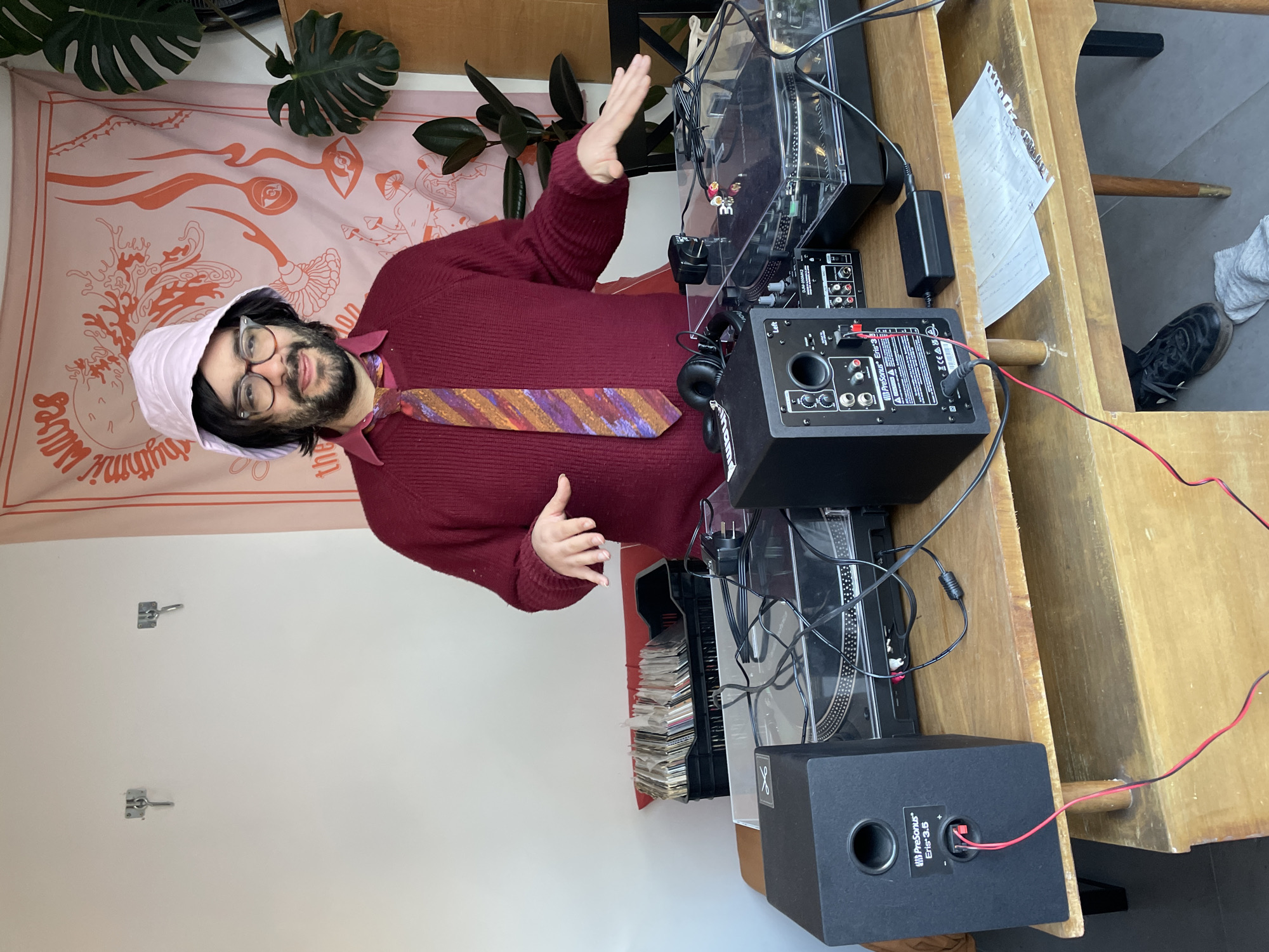 A man wearing glasses, a pink hat, and a red sweater with a colorful tie is standing behind a turntable set up. The setup includes two turntables, a mixer, and two black speakers. The equipment is placed on a wooden table, and there is a stack of records to the left of the turntable. Behind the man is a wall with two hooks, and a tapestry with an image of mushrooms and eyes. To the right of the setup is a section of wall, part of a pair of shoes and some papers. There is no immediate evidence suggesting that the scene is located in Buenos Aires, Argentina.