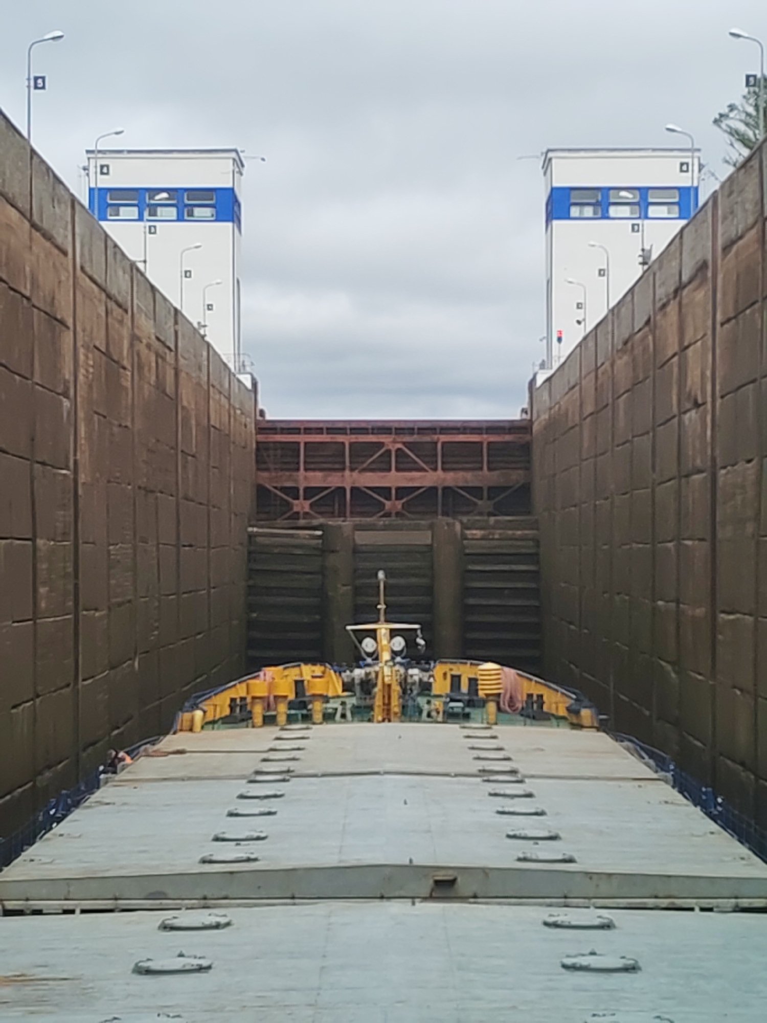 The image depicts a barge within the lock of the Volga-Baltic Waterway in Devyatiny, Russia. The barge, with a gray deck and yellow machinery, is situated between two tall, brown concrete walls. Two white towers with blue horizontal stripes stand on either side of the lock. The lock's gate is visible in the background, made of brown steel. The sky is overcast with gray clouds.