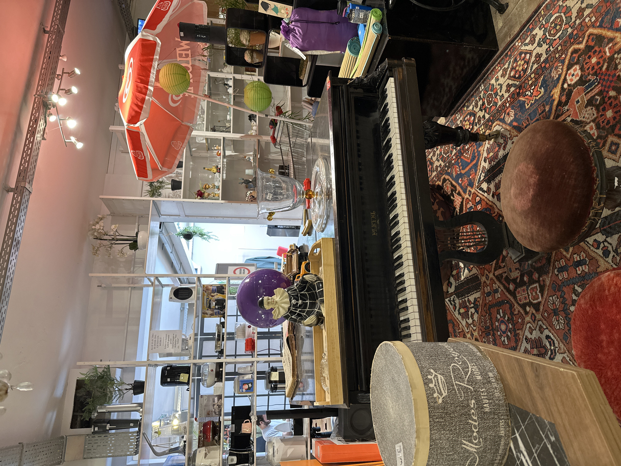 The image shows the interior of a cluttered shop with shelves filled with various items, including kitchenware, decorative objects, and plants. In the foreground, a grand piano is positioned on a patterned rug. On top of the piano, there is a ceramic clown figurine, a large purple ball, and a tray with decorative items. To the left of the piano, a round grey container with a crown and text that reads "Modes Royal HAUTES MODES ANVERS" is visible. A man is partially visible in the background, standing near a shelf. An open umbrella with the logo "TCHIBO" is displayed above the piano. The environment appears to be a retail space, possibly a second-hand shop or a boutique, located in Bruges, Belgium. The lighting is a mix of natural and artificial sources, with spotlights on the ceiling illuminating the scene.
