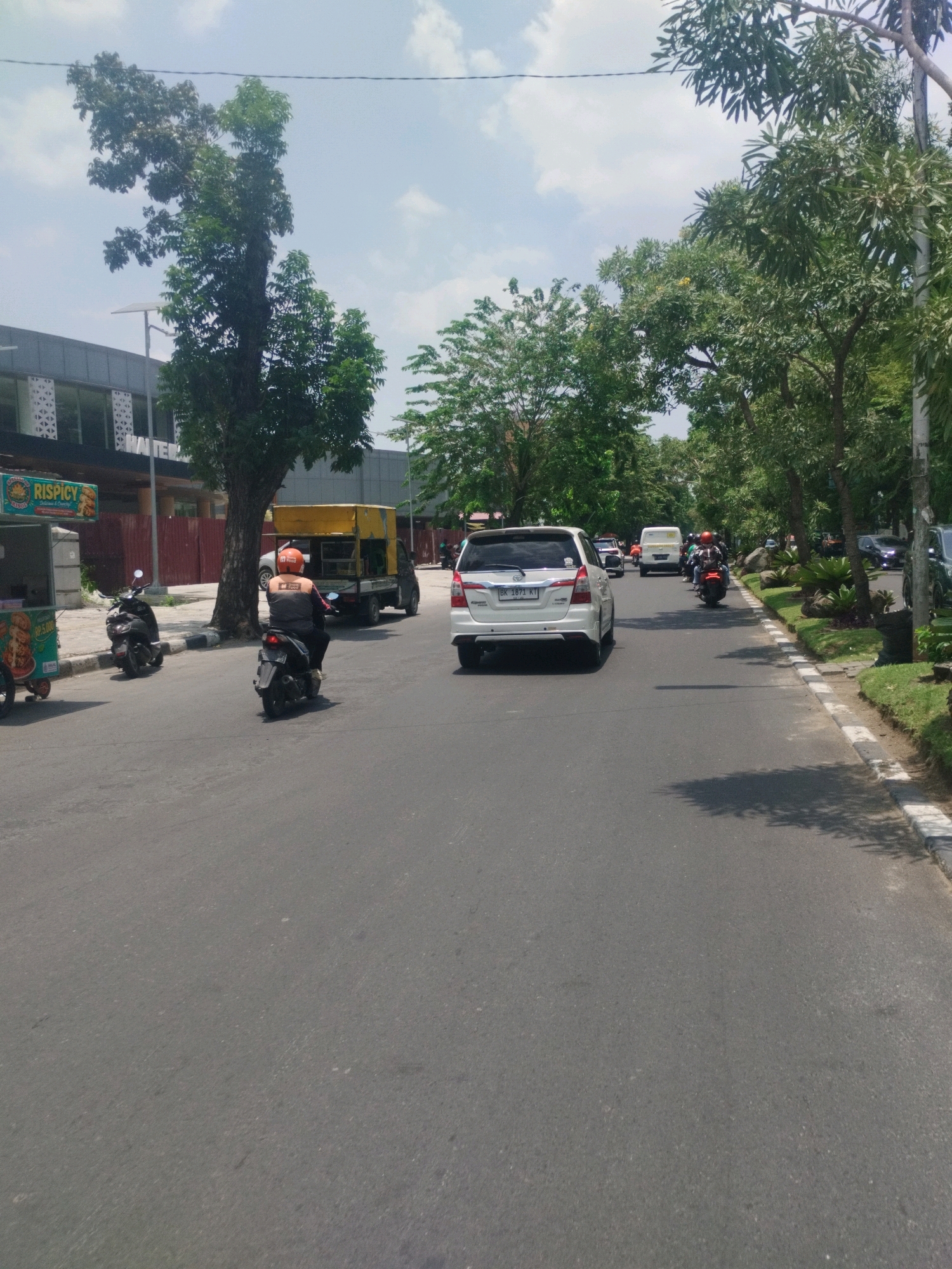 A bright, sunny day is depicted on a paved street in the city of Medan, Indonesia. The street is lined with lush green trees on the right side, casting shadows on the asphalt. On the left side of the street, a modern building with large windows and patterned facade is visible, along with a food cart selling "RISPY SPICY" items. Several vehicles are present on the road, including a white Toyota Innova, a yellow truck, and multiple motorcycles. One motorcycle rider, wearing an orange helmet and a grey jacket with orange accents, is in the foreground, moving away from the camera. Other vehicles and riders are further down the street, indicating traffic. The sky is partly cloudy, with scattered white clouds against a light blue backdrop. The overall scene suggests a typical day in a developed urban area of Medan.