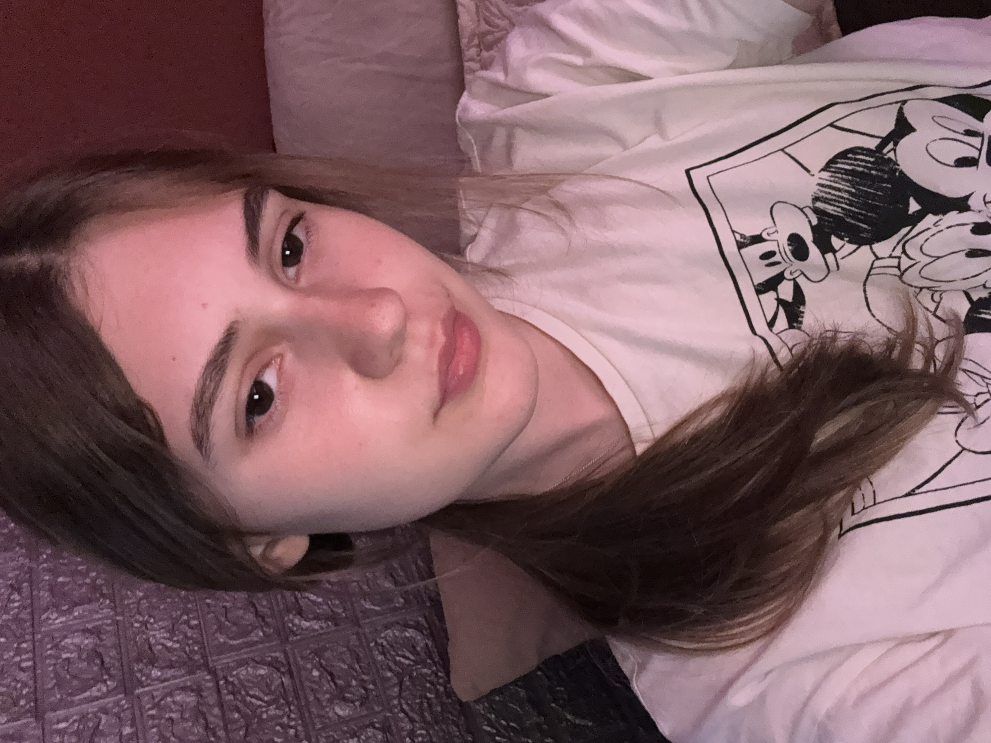 The image displays a young woman lying down indoors in Panagyurishte, Bulgaria, looking directly at the camera. She has medium-length brown hair, dark eyes, and a neutral, relaxed expression. She is wearing a light-colored t-shirt that features a prominent black outline graphic of Mickey Mouse and other cartoon characters in a comic strip style.
Her head is slightly tilted, resting on what appears to be a light-colored pillow or bedding. To the upper left, a dark red wall or headboard is visible. Below her, on the left side of the frame, there is a dark, textured surface with an embossed, repeating pattern. The lighting in the scene is soft and warm, casting a gentle, slightly pinkish glow, suggesting an indoor environment, possibly in the evening or a dimly lit room. A thin, silver-colored chain is faintly visible around her neck.