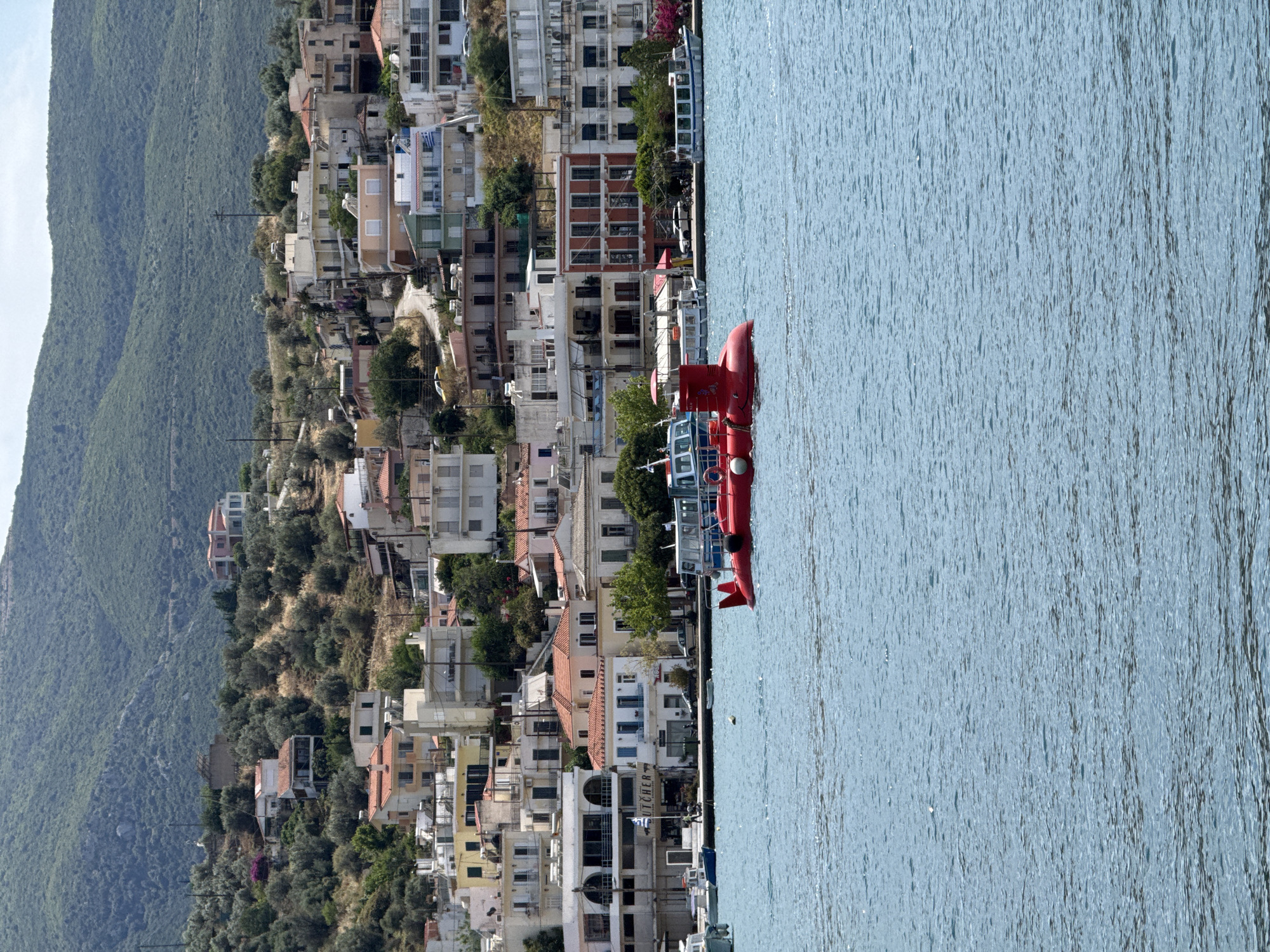 The image depicts a hillside urban landscape in Poros, Greece, adjacent to the water. On the left, a lush green mountain slopes down to a dense cluster of buildings characterized by white and earth-toned structures with red-tiled roofs. The buildings are tightly packed together, typical of a hillside town.
On the right, the calm blue water extends toward the horizon, reflecting the sky. Several red and white boats are docked along the waterfront. The town's buildings are reflected on the water’s surface, creating an interplay of reality and reflection.