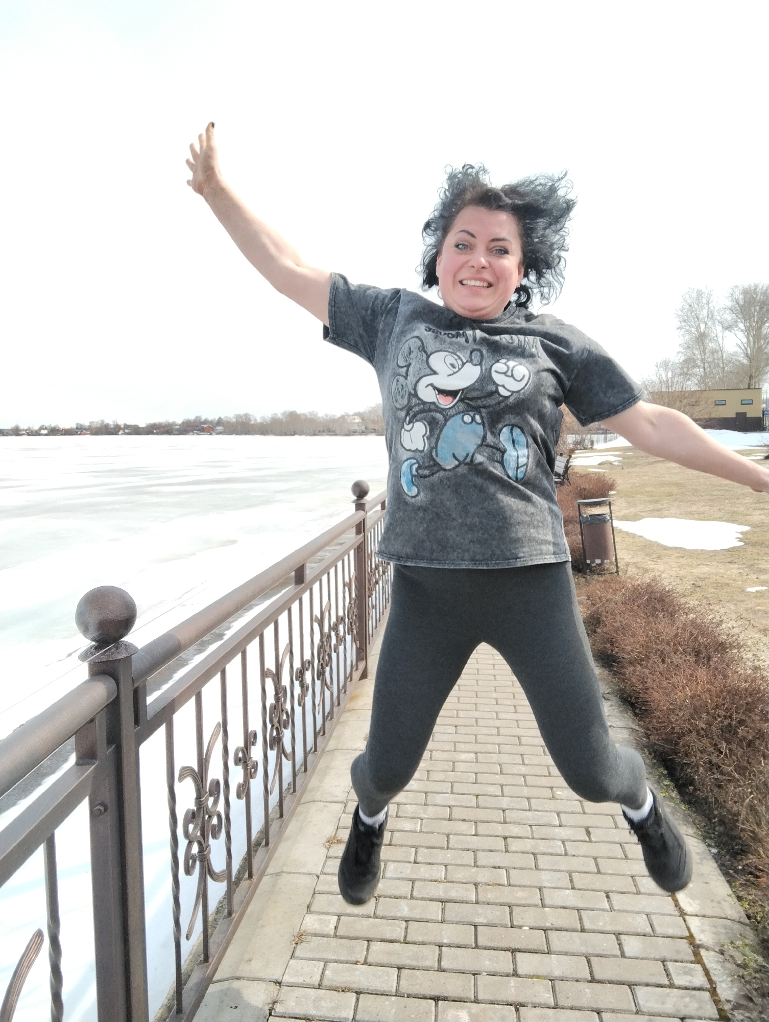 A woman is jumping on a brick walkway in Votkinsk, Russia. The woman has dark hair, is wearing a gray t-shirt with a Mickey Mouse image on the front, dark gray leggings, and black shoes. She has one arm raised above her head and the other extended to the side. A metal railing is on the left, separating the walkway from a frozen body of water covered in snow. On the right is a grassy area with patches of snow and brown bushes. In the background are trees, buildings, and a light sky.