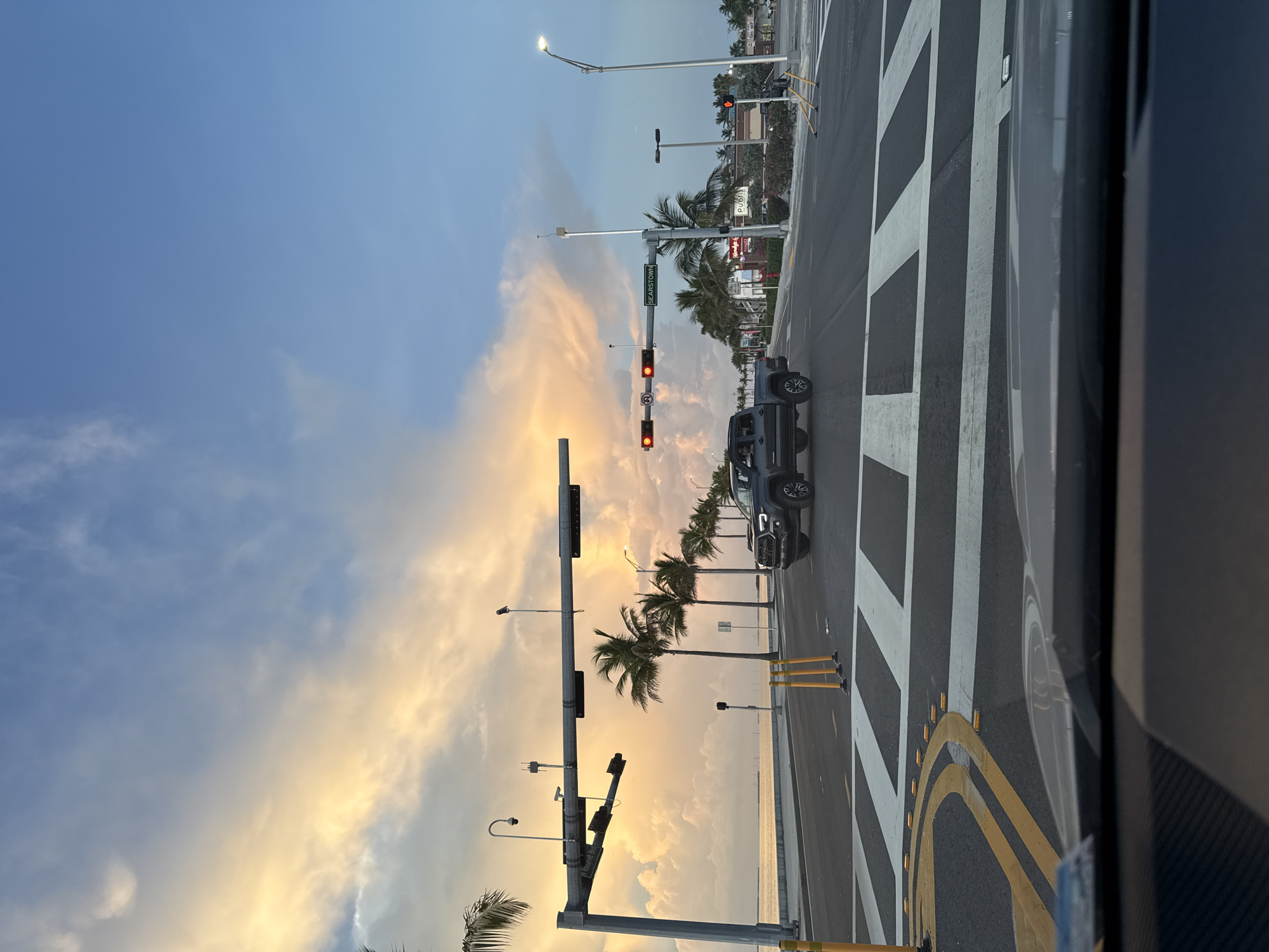 A dark gray Toyota Tacoma pickup truck drives through an intersection in Key West, Florida, USA. The truck is traveling east on Roosevelt Boulevard. The sky above is a mix of blue and dramatic, yellow-tinged clouds, indicative of sunset. Palm trees line the north side of the boulevard, and a body of water is visible beyond them. Traffic lights are red in both directions, and a street sign reads "SEARSTOWN". The foreground shows the dashboard and windshield of a vehicle that is stopped at the intersection, facing east. The road features white pedestrian crosswalk stripes and yellow lane markings.