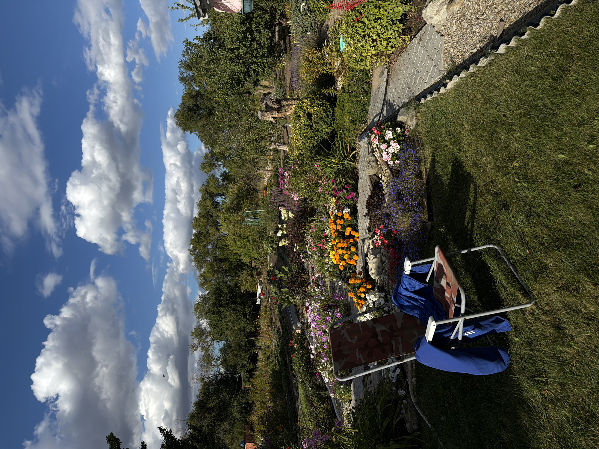 A well-maintained garden in Kamyshin, Russia, features a variety of blooming flowers, lush green trees, and a cloudy blue sky. A camouflage-patterned folding chair with a blue jacket draped over it sits on a patch of manicured grass in the foreground. A stone pathway winds through the garden, bordered by gravel and more vibrant flowers. In the background, more trees and plants create a natural backdrop, with a wooden stump adding a rustic touch. The overall impression is one of a peaceful, cultivated outdoor space.