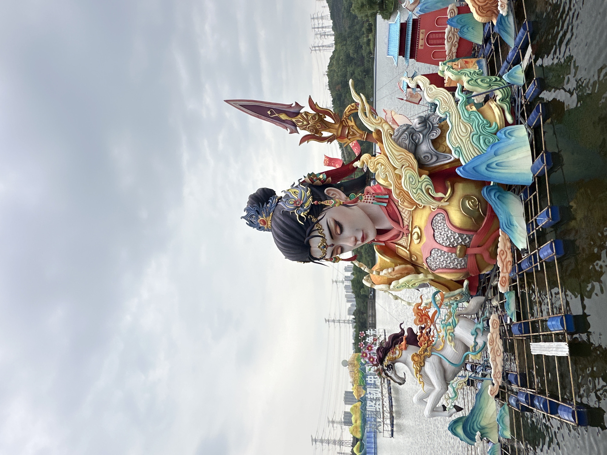 The image depicts a large, ornate float on the Liangjiang River in China. The float features a figure of a female deity with elaborate headwear and a golden breastplate. She holds a spear pointing upwards. Below the deity are several horse figures also decorated in bright colors. The float is constructed on a base of blue barrels and metal supports that are partially submerged in the water. In the background, trees and buildings line the riverbank, with power lines visible in the distance. The sky is overcast.