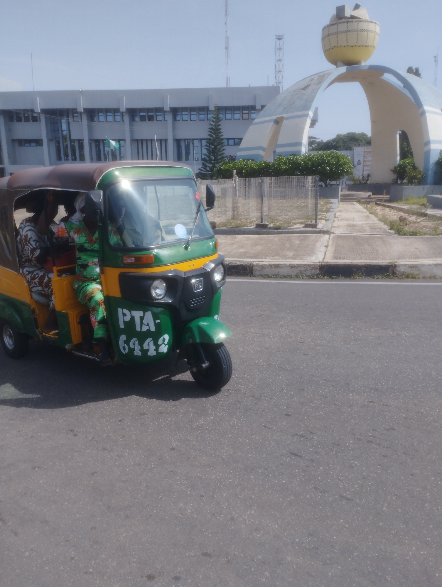 An eye-level, full shot captures a three-wheeled motorized rickshaw, commonly known as an auto-rickshaw or Keke Napep, driving on a paved road under a clear sky. The auto-rickshaw, painted in a distinct green, yellow, and orange livery, has "PTA-6442" painted in white on its front side panel. Three individuals are seated inside the vehicle; one driver in the front and two passengers in the back, all appearing to be adults. The passengers are wearing traditional attire.
In the background, a modern, grey, two-story building with numerous windows is visible. To the right of the building, a large, abstract architectural structure, primarily white with blue accents, arches over a pathway. This landmark is topped with a yellow, globe-like sculpture adorned with a decorative pattern. In front of the building and landmark, there are trimmed green bushes and a grey chain-link fence. The environment suggests an urban setting, likely a public space or a governmental area. The overall lighting indicates it is daytime, with a slight glare on the auto-rickshaw's windshield. The specific location is implied to be Jos, Nigeria, based on the presence of the architectural landmark and the type of vehicle.