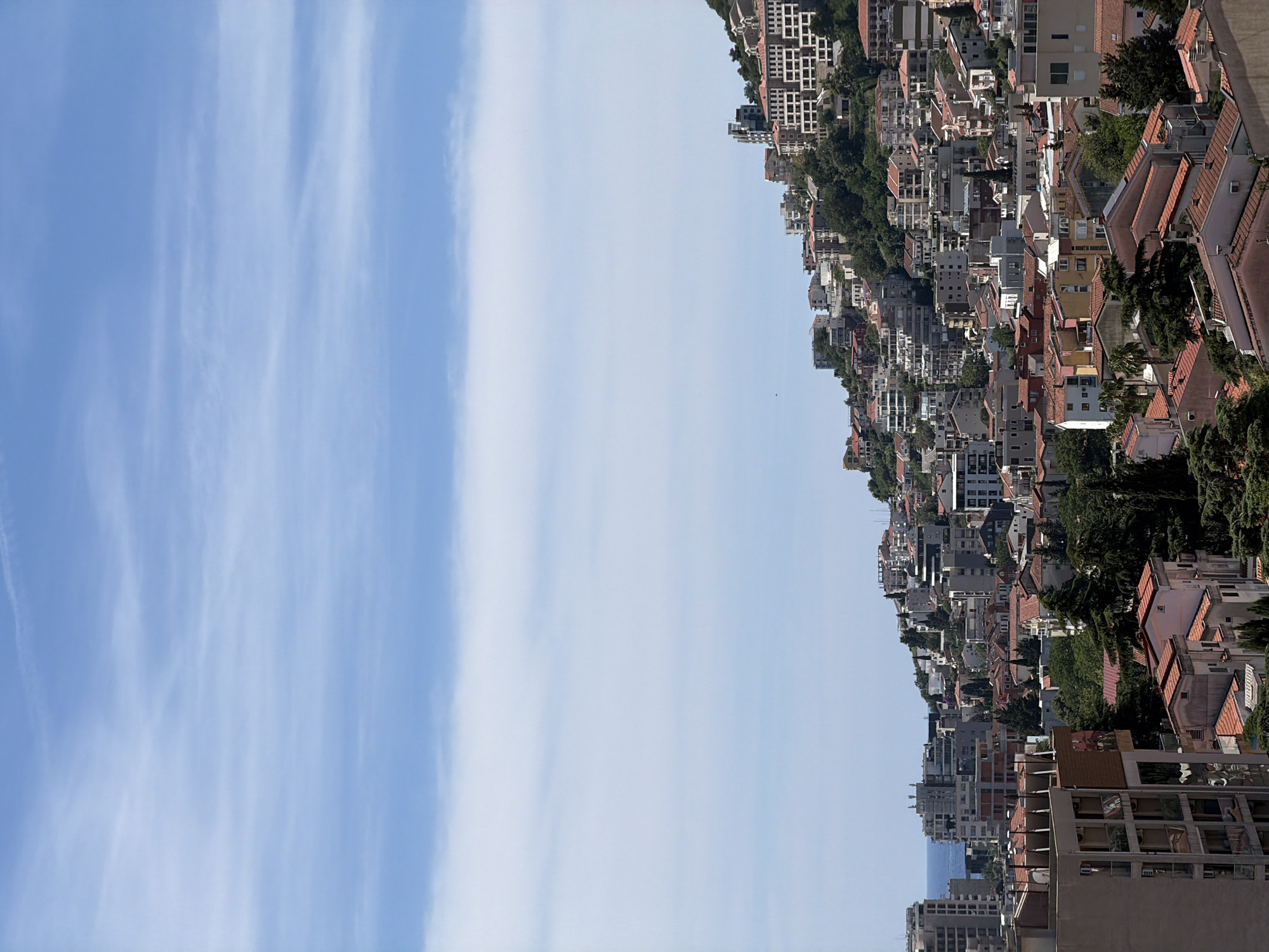 The image displays a densely populated hillside in Budva, Montenegro, overlooking the Adriatic Sea. The scene is dominated by a vast expanse of blue sky with scattered, wispy white clouds. Below the sky, a gradient of blues transitions to the horizon where the sea is faintly visible. The hillside is covered with numerous buildings, ranging from multi-story apartment complexes and hotels to smaller houses, all constructed with a mix of white, beige, yellow, and red facades. Many buildings feature terracotta-tiled roofs. Lush green trees and foliage are interspersed throughout the urban landscape, particularly on the steeper parts of the hill. The architecture suggests a Mediterranean style, with balconies and varied building heights creating a textured, layered appearance. In the foreground, several buildings are more clearly defined, revealing details of their construction and residential nature. The overall impression is one of a vibrant coastal city built on a hilly terrain. No people are visible in the image.