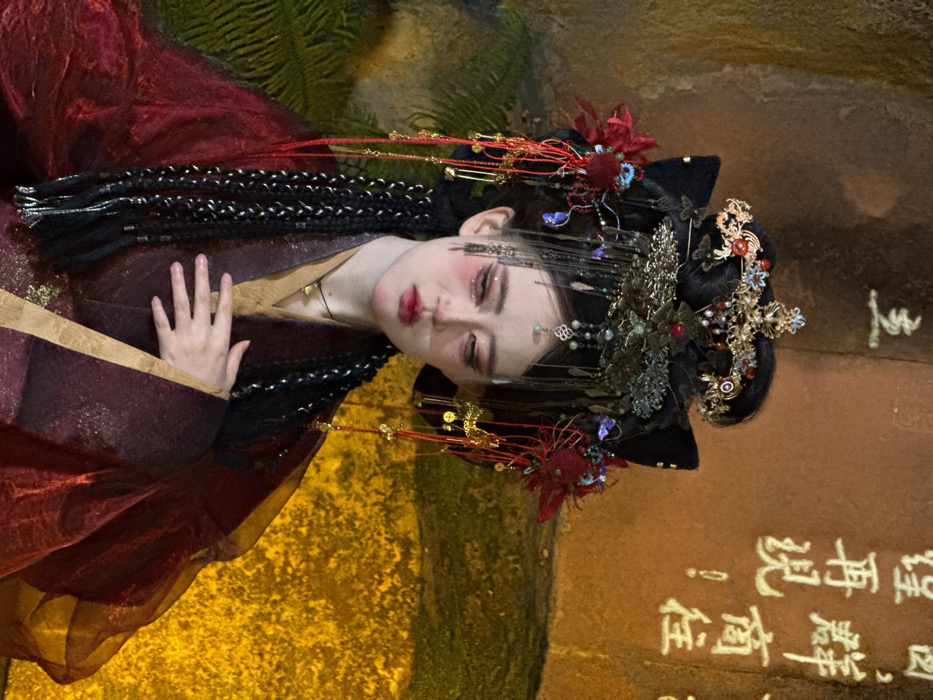 A close-up, chest-up image depicts an individual in elaborate traditional Chinese attire, positioned against an illuminated, textured wall. The individual has a pale complexion, prominent theatrical red makeup under the eyes and on the cheeks, and red lipstick. Their black hair is styled intricately, featuring long braids interwoven with silver strands extending over the shoulders. A complex, multi-layered headpiece adorns the head, incorporating black velvet structures resembling cat ears, gold-toned filigree, red and blue embellishments, and multiple dangling chains with small gold-colored coin-like charms. The clothing consists of a gold/beige inner garment beneath a translucent deep red outer robe with subtle patterning. A black cord necklace with a small rectangular gold pendant is visible. The individual's gaze is directed downwards, and their right hand rests open-palmed on the chest area. The background features a rough, vertically textured wall or rock face, illuminated by a warm, yellowish-orange light. White Chinese characters are visible on the wall, with fragments such as "立", "群商往", and "望再現!" discernible. Green foliage is partially visible in the lower left background. The scene is set in Yuzhong District, China.