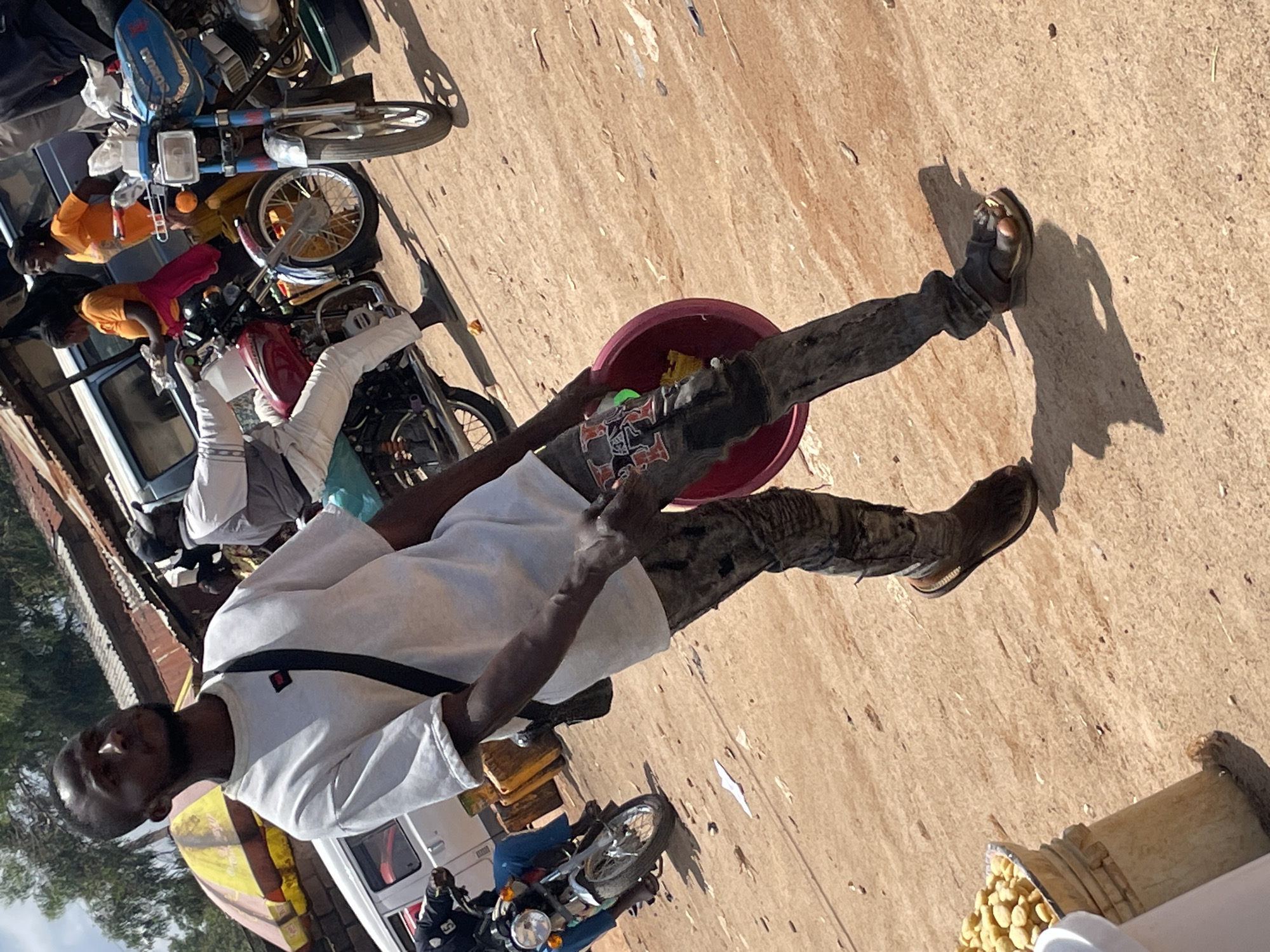 The image captures a street scene in Wang, Nigeria, with a focus on a young man in the foreground. He is wearing a white t-shirt and dark, heavily distressed jeans. A black strap from a bag crosses his chest. In his right hand, he holds a reddish-pink basin containing what appear to be yellow food items, possibly snacks. His left hand is near his hip. He is wearing dark sandals, and his left foot is visible.
The background reveals a bustling environment with several motorcycles. One motorcycle, partially obscured, is in the lower left. Further back, another motorcycle is prominent, a blue and white Suzuki, with its front wheel turned to the right. Behind the motorcycles, various structures and vehicles are visible, including parts of vans or trucks. There are also people, including a woman in an orange t-shirt and another in a white shirt, interacting or present in the scene. Overhead, a yellow and red umbrella is visible, suggesting a market or roadside vendor area. The ground is a dusty, sandy surface typical of many Nigerian roads. The overall impression is that of a lively, somewhat chaotic, outdoor marketplace or transportation hub.
