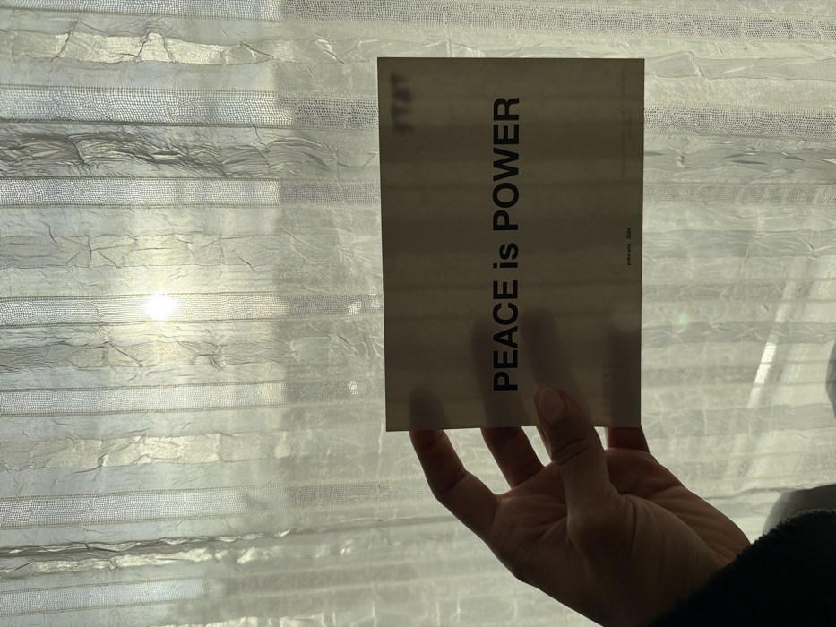 An adult human hand, appearing to be a right hand, holds a rectangular, light-toned card horizontally in the foreground. The card displays the prominent black text "PEACE is POWER" in a sans-serif font, with "yoko ono 2024" printed in smaller text below. A partial word, possibly "STAY," is visible in the upper right corner of the card. The hand's thumb secures the card from the top, while fingers are positioned underneath. The individual's dark clothing is visible at the lower left edge.
The background consists of a sheer, light-colored curtain, exhibiting vertical striped patterns and a wrinkled texture, partially obscuring an exterior view. A brilliant, circular light source, consistent with the sun, is visible through the curtain, centered in the upper background, producing a strong glare and backlighting the scene. Faint, dark silhouettes of distant structures, resembling urban buildings, are discernible through the curtain beyond the bright light. The scene is located in Champs-sur-Marne, France.