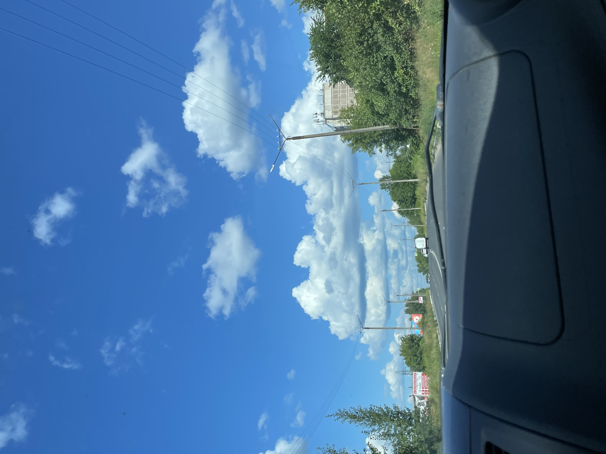 The image is a point-of-view shot from inside a vehicle, looking out at a road on a sunny day. The sky is bright blue with scattered white cumulus clouds. Power lines are visible stretching across the frame. On the right side of the road, there are trees and a multi-story building. On the left side of the road, there are more trees and a billboard. The road curves to the right in the distance, and other vehicles are visible. The dashboard of the vehicle is in the foreground. The scene is consistent with a suburban or semi-rural area.