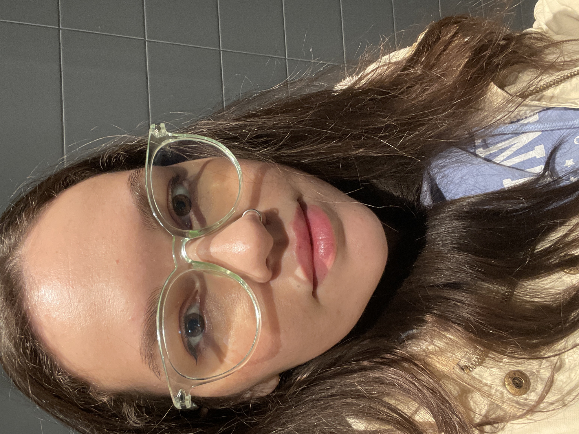 A close-up, angled portrait features a young person with long, dark brown hair, likely taken indoors in Izhevsk, Russia. The subject's head is tilted to the left, showing their face in a slightly upward gaze. They wear light green-framed, round glasses through which their brown eyes are visible. A silver nose ring adorns the left nostril, and their lips are a soft pink shade.
The individual's hair frames their face and extends around their shoulders, appearing somewhat disheveled. They are dressed in a light blue garment, possibly a hoodie or t-shirt, which features white graphics including stars and partially visible letters resembling 'U' and 'S' or 'A'. Over this, a light-colored (beige or cream) jacket with a zipper and visible snap buttons is worn. The background consists of a dark gray tiled wall. The lighting suggests an indoor setting, possibly from an overhead source, illuminating the face. The expression is neutral.