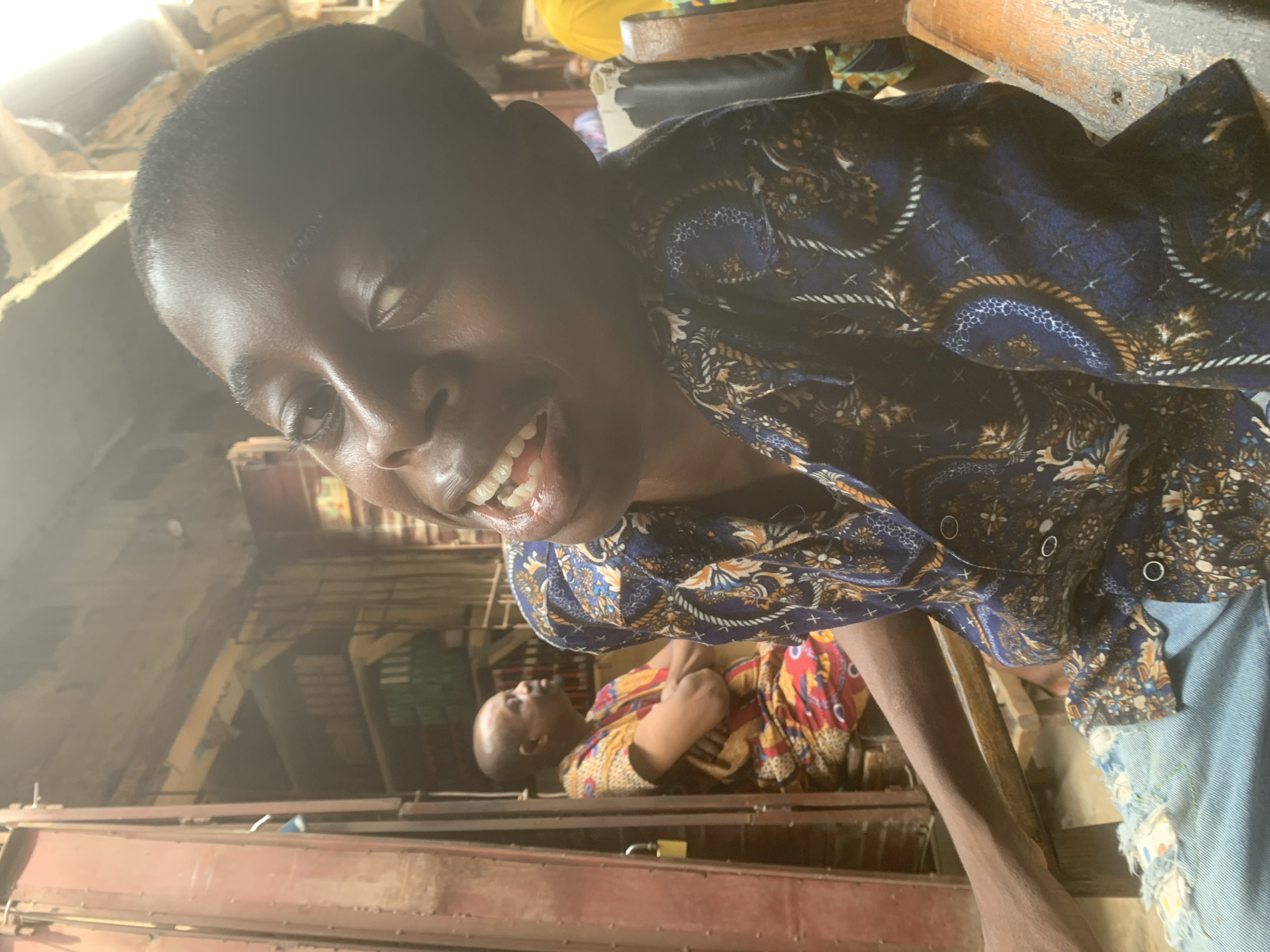 This indoor photograph, taken in Fegge, Nigeria, features two individuals. The primary subject, a young person with dark skin and short hair, is captured in a close-up, smiling broadly and looking directly into the camera. They are dressed in a dark blue short-sleeved shirt adorned with intricate patterns in gold, white, and lighter blue, along with light blue denim shorts or trousers that show some distressed detailing.
In the mid-ground, another person, also appearing young with dark skin and short hair, is visible lying down, clad in a brightly colored, patterned garment featuring shades of red, yellow, and black. The background suggests an interior space, possibly a dwelling or a storage area, with various wooden and metal structures, some appearing as shelving or beams. A padlock is visible on a metal bar in the lower left, hinting at a secure or enclosed area.
The scene is well-lit by natural light, casting highlights on the main subject's face and indicating it is daytime. The main subject's wide smile conveys a clear sense of happiness and engagement. There is no visible text in the image.