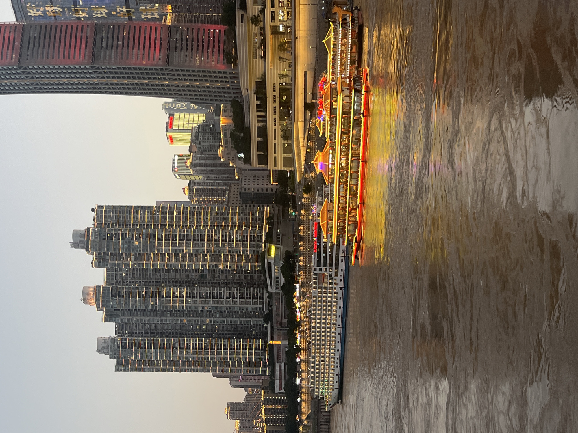 The image captures a dusk scene along the Jialing River in Nan'an District, Chongqing, China. Dominating the background are numerous modern skyscrapers, many of which are illuminated with warm, yellow lights, creating a vibrant cityscape. In the middle ground, a large white cruise ship is docked or moving slowly, its windows also aglow with interior lights. To its right, a smaller, elaborately decorated boat, featuring a red and yellow exterior with illuminated details, is visible. The Jialing River itself is a muddy brown, reflecting the lights from the city and the boats, creating a shimmering effect on the water's surface. The sky is a pale, hazy grey, typical of twilight conditions. No individuals are clearly discernible in the image, and the focus is on the architectural and nautical elements of the scene.