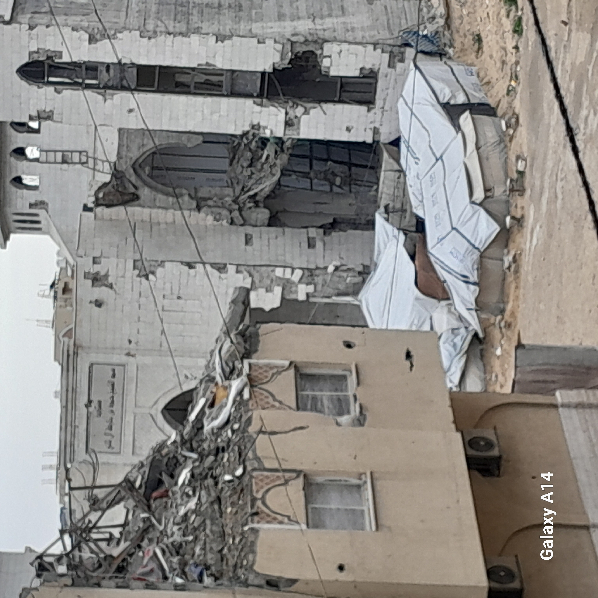 The image depicts a severely damaged building in Khan Yunis, Palestinian Territory. The structure has sustained significant damage, with large portions of the walls and roof collapsed. Debris is scattered around the building. Adjacent to the damaged building is another structure, partially visible, with visible windows and air conditioning units. A pile of white objects are located to the right of the damaged building.
