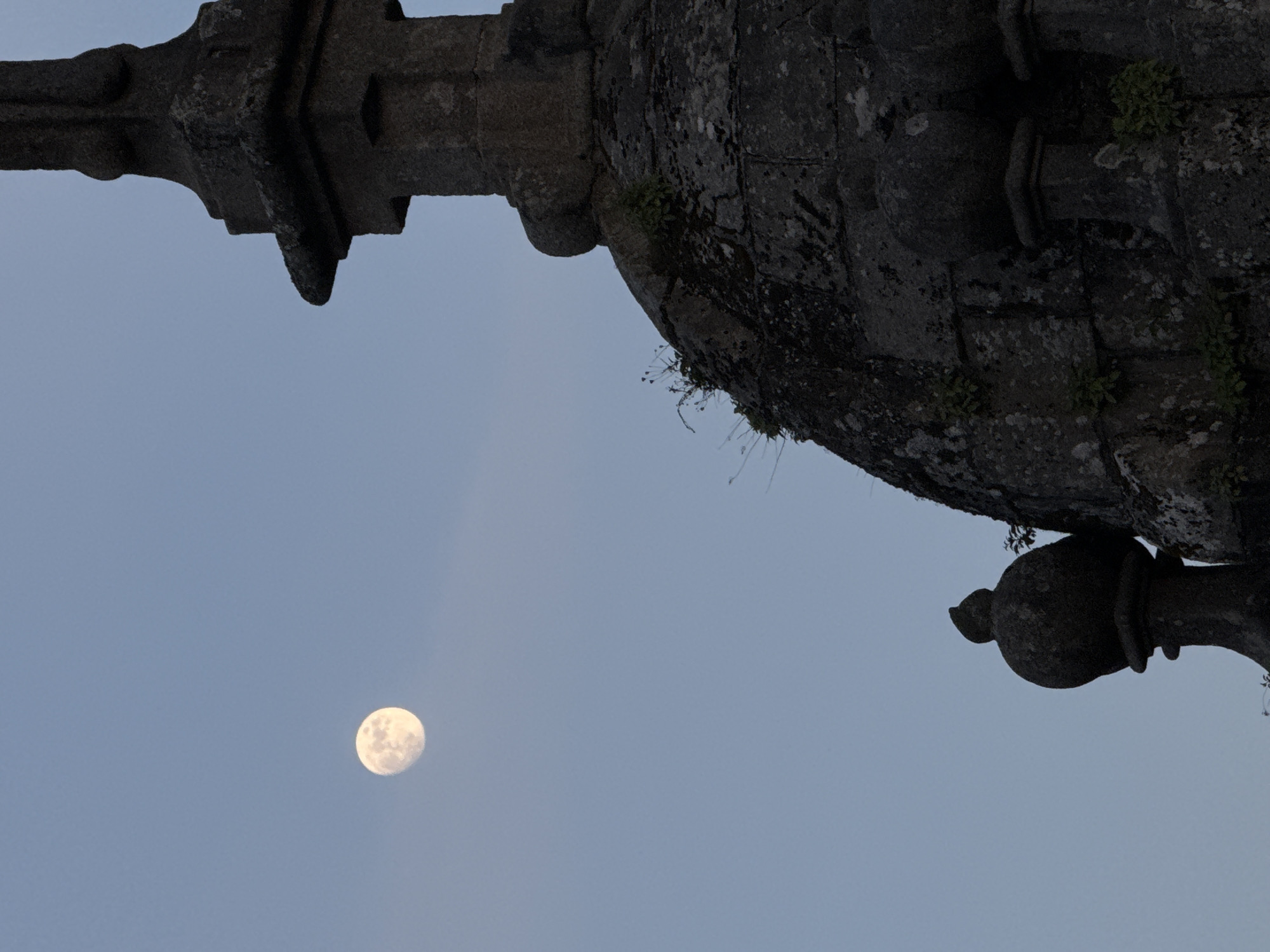 The image depicts a full moon in the sky, observed through the architectural features of a building in Santiago De Compostela, Spain. The structure is made of stone and shows signs of weathering and age. There are some small plants growing on the stone. The building is a historical monument, likely part of the cathedral or another significant structure in the city. No people are visible.