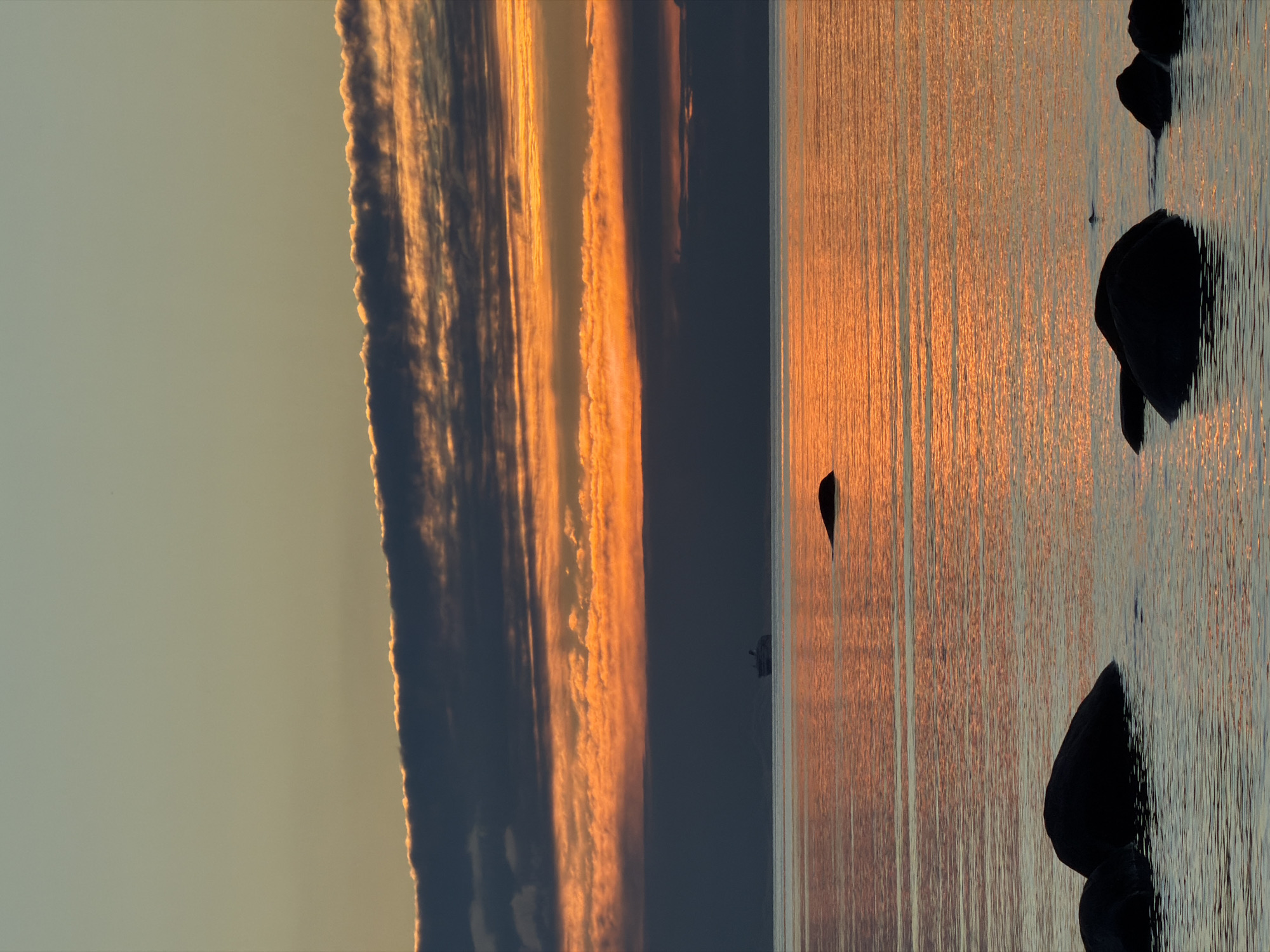 The image depicts a serene seascape at dusk in Haabneeme alevik, Estonia. The sky is a canvas of warm, diffused light, transitioning from a pale gold near the horizon to a soft blue overhead. A band of dark, striated clouds stretches across the middle of the frame, tinged with fiery orange and yellow where they catch the last rays of the sun. Below the clouds, a thin layer of haze softens the distant horizon.
The sea is calm, reflecting the vibrant colors of the sky in shimmering horizontal streaks. A large ship, possibly a ferry or cargo vessel, is visible as a dark silhouette on the water in the distance, almost merging with the haze. In the foreground, several dark, rounded rocks emerge from the water, their shapes softened by the twilight. One solitary rock breaks the surface further out on the water, a small, dark shape against the glowing reflection. The overall impression is one of tranquility and natural beauty as day gives way to night.