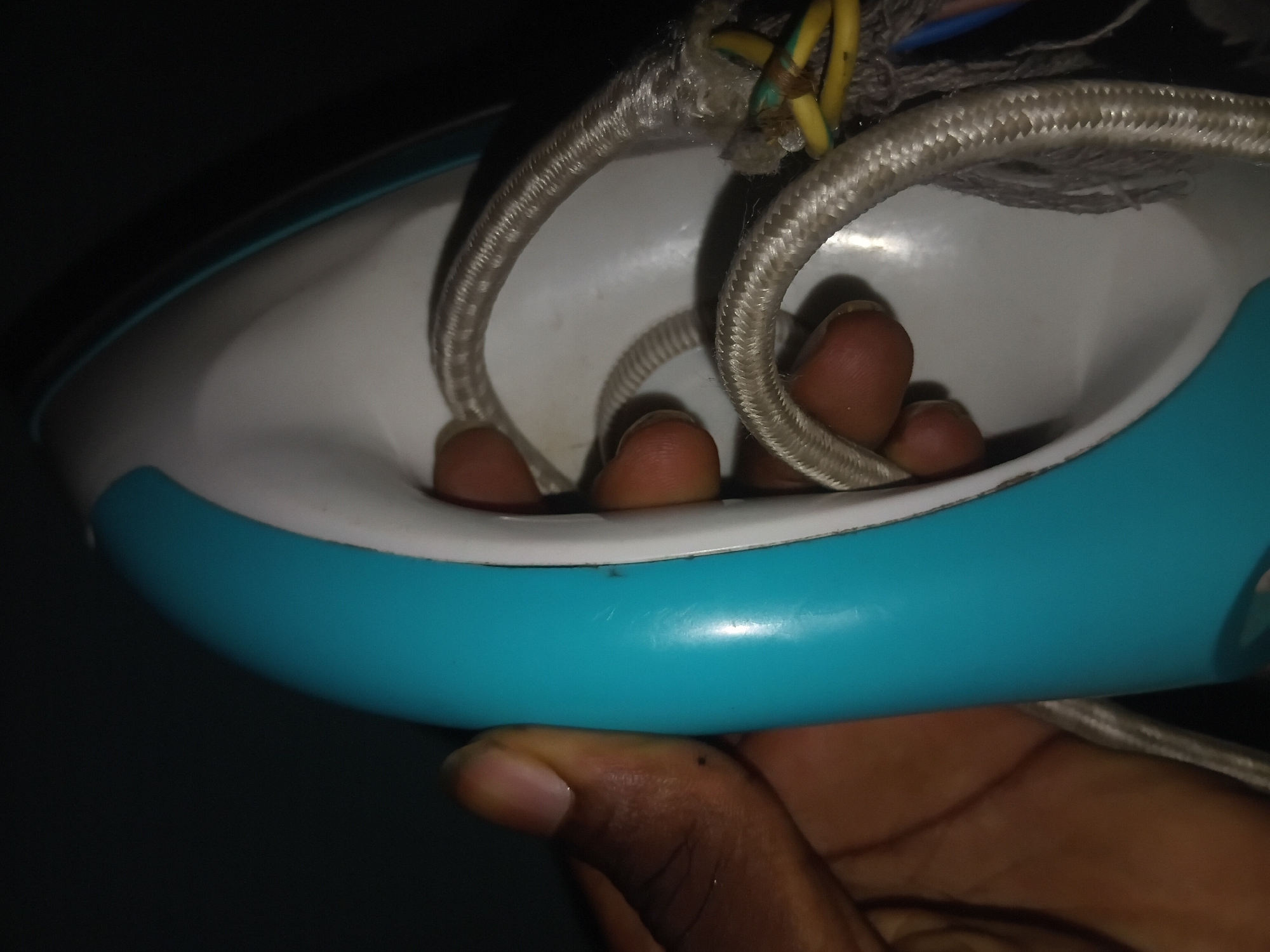 A close-up, low-angle, side view of a person's hands holding a white and turquoise iron. The iron's power cord, which is frayed and exposes yellow and blue wires, is visible in the foreground. The background is dark and out of focus. The image appears to be taken indoors in Jalingo, Nigeria, possibly in a residential setting due to the domestic appliance and the visible electrical wiring. The lighting suggests it may be evening or a dimly lit room.