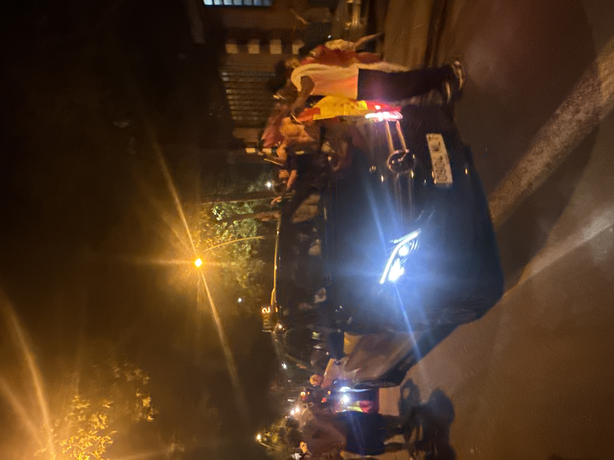 An outdoor, nighttime, street scene in Barcelona, Spain, shows a black Mercedes-Benz sedan with its headlights illuminated and reflecting off the wet pavement. The license plate, "0904 MLT," is visible. Several people are gathered around the car, some appearing to be celebrating. One individual on the right is wearing a white t-shirt and dark pants, holding a yellow and red flag. To the left of the car, a person with their back to the camera is wearing a dark vest or shirt and shorts, and appears to be holding something. Another individual in the background on the far left is wearing a light-colored top and dark bottoms. A taxi sign reading "TAXI 2" is mounted on the roof of a vehicle behind the Mercedes. Trees with illuminated foliage are visible on the left side of the frame, and a building with barred windows is on the right. Streetlights cast an orange glow, creating lens flares in the image. The overall atmosphere appears to be festive.