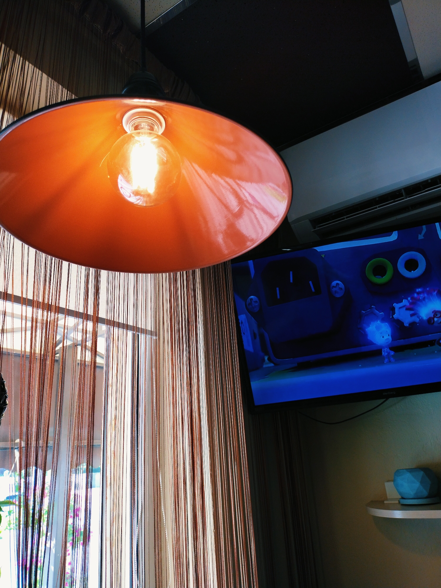 The image captures an indoor scene, likely a room in Horishni Plavni, Ukraine. The foreground is dominated by a large, bright orange lampshade hanging from the ceiling, with an Edison-style lightbulb glowing warmly within it. Behind the lampshade, a window is visible, covered by thin, vertical, copper-colored strands that create a semi-transparent curtain.
To the right of the lampshade and curtain, a flat-screen television is mounted on the wall. The television displays a cartoon, featuring animated characters in a blue-hued environment. Above the television, an air conditioning unit is mounted. Further to the right, a small, light-colored shelf is attached to the wall, holding a geometrically shaped teal-colored object and a small white item. The wall itself is a muted beige color. The overall lighting suggests either late afternoon or early evening, with artificial light from the lamp and television contributing to the ambiance.
