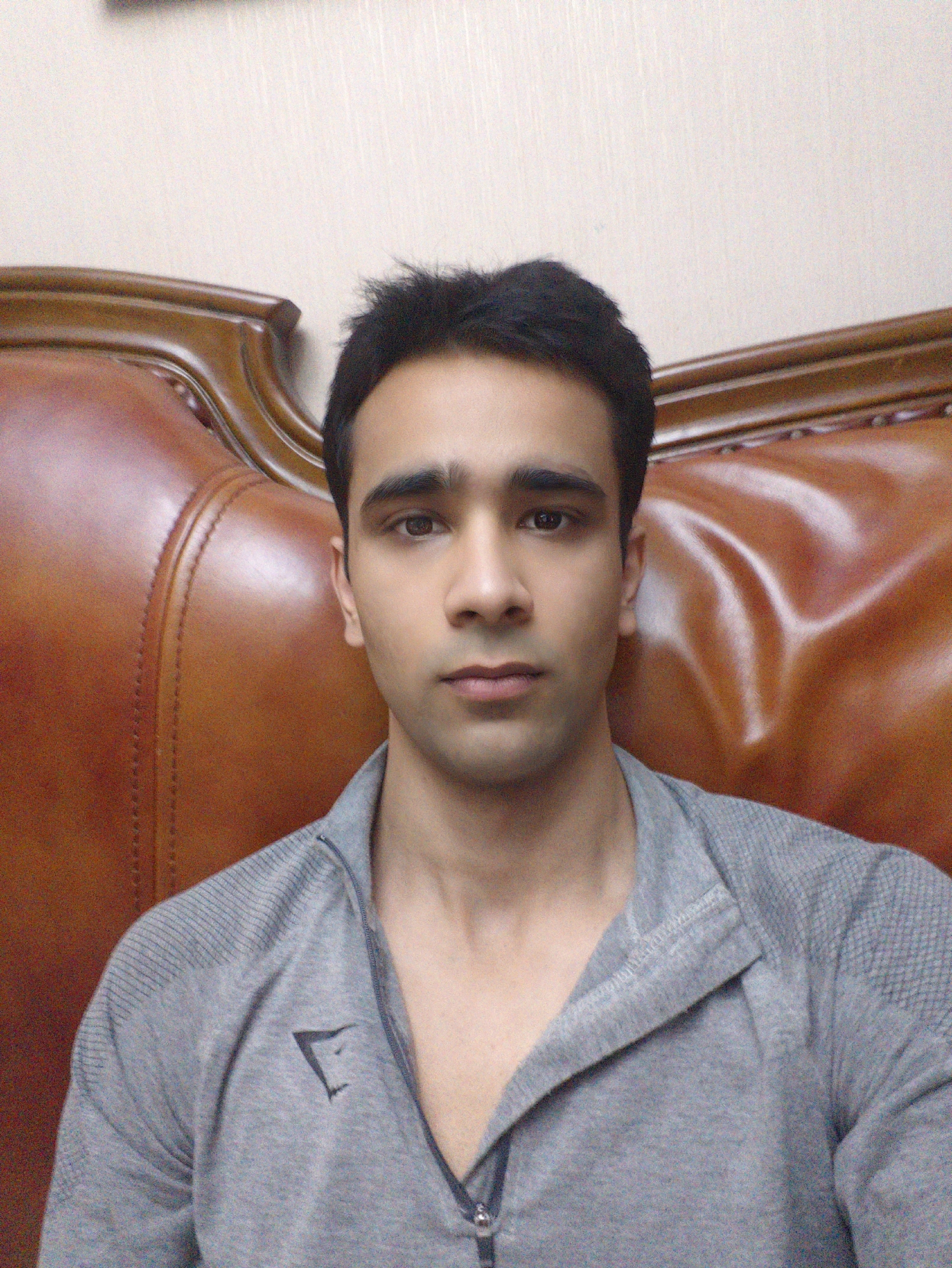 A man with dark hair and brown eyes is seated in a brown leather chair. He is wearing a grey zip-up shirt with a small logo on the left side. The background is a plain, light-colored wall. There is no information to determine the specific location in Chongqing High-tech Zone, China.