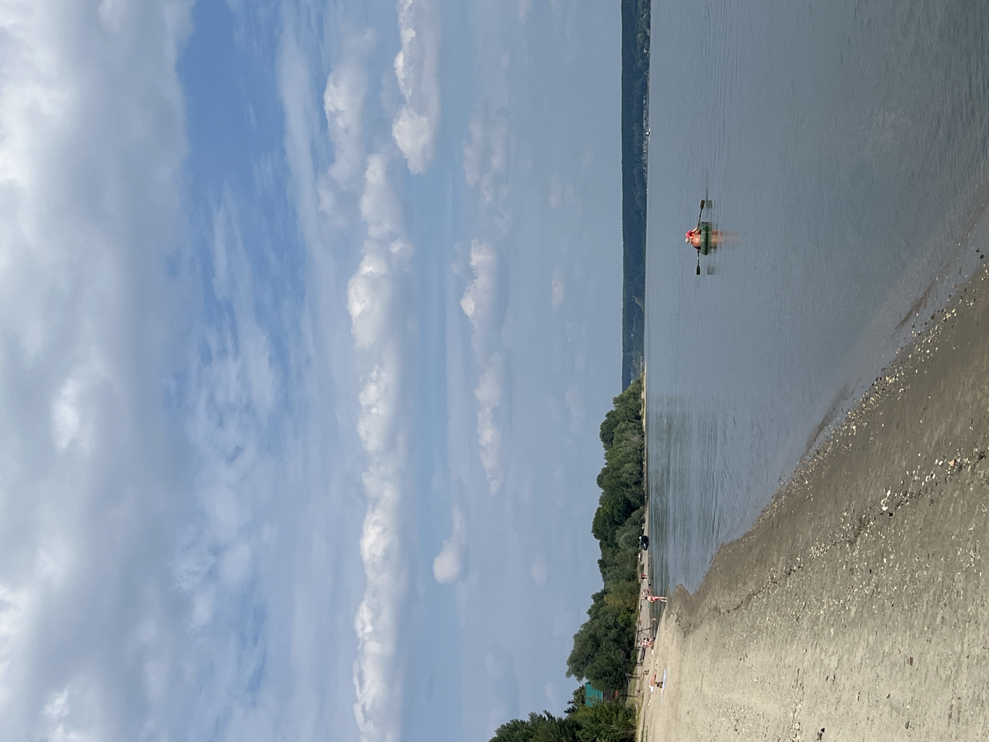 A wide, calm river flows from right to left, reflecting the overcast sky. On the left bank, a sandy beach stretches into the distance, dotted with a few people sunbathing and walking. The beach is bordered by a dense line of green trees and some buildings. Further down the river, a small group of people are in an inflatable boat, with two individuals actively rowing. The opposite bank is covered by a dark line of trees, suggesting a vast, undeveloped landscape. The overall impression is of a serene, summery day by the water in the village of Mochishche, Russia.