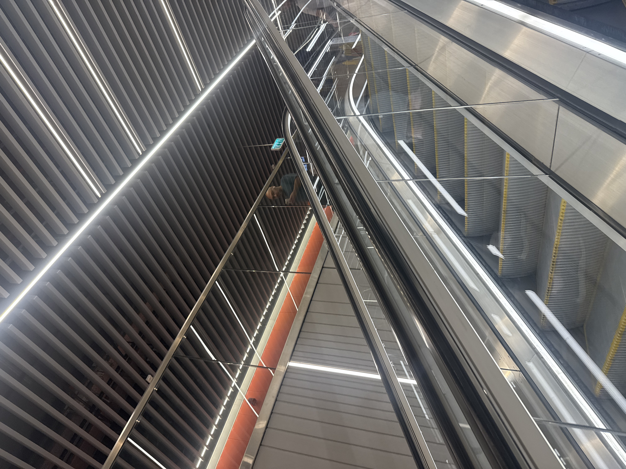 This image presents a modern architectural interior, likely a station or large building in Ramat Gan, Israel. The scene is dominated by strong diagonal lines and reflective surfaces. On the right, two escalators ascend, featuring metallic handrails, glass side panels, and steps with yellow-edged treads. The glass and polished metal surfaces reflect the surrounding structure.
To the left, a prominent slatted wall extends upwards, composed of numerous grey horizontal panels. Bright, linear LED lights are integrated diagonally within these slats, creating luminous stripes. A triangular section between the slatted wall and the escalators features a lighter grey surface, bordered by a vibrant orange strip on its left side, also adorned with linear lighting.
A single person is partially visible in the middle-left, reflected and somewhat obscured by the structure. They appear to be wearing a dark top and are looking downwards, suggesting they might be on a landing or another escalator level. Their presence indicates public access and use of the facility.
The image is captured from a low angle looking up, highlighting the height and contemporary design. The artificial lighting from the integrated LEDs creates a bright, clean, and futuristic aesthetic. A small blue rectangular sign with illegible white text can be seen near the individual. The overall impression is one of sleek design, order, and efficient public transit infrastructure.