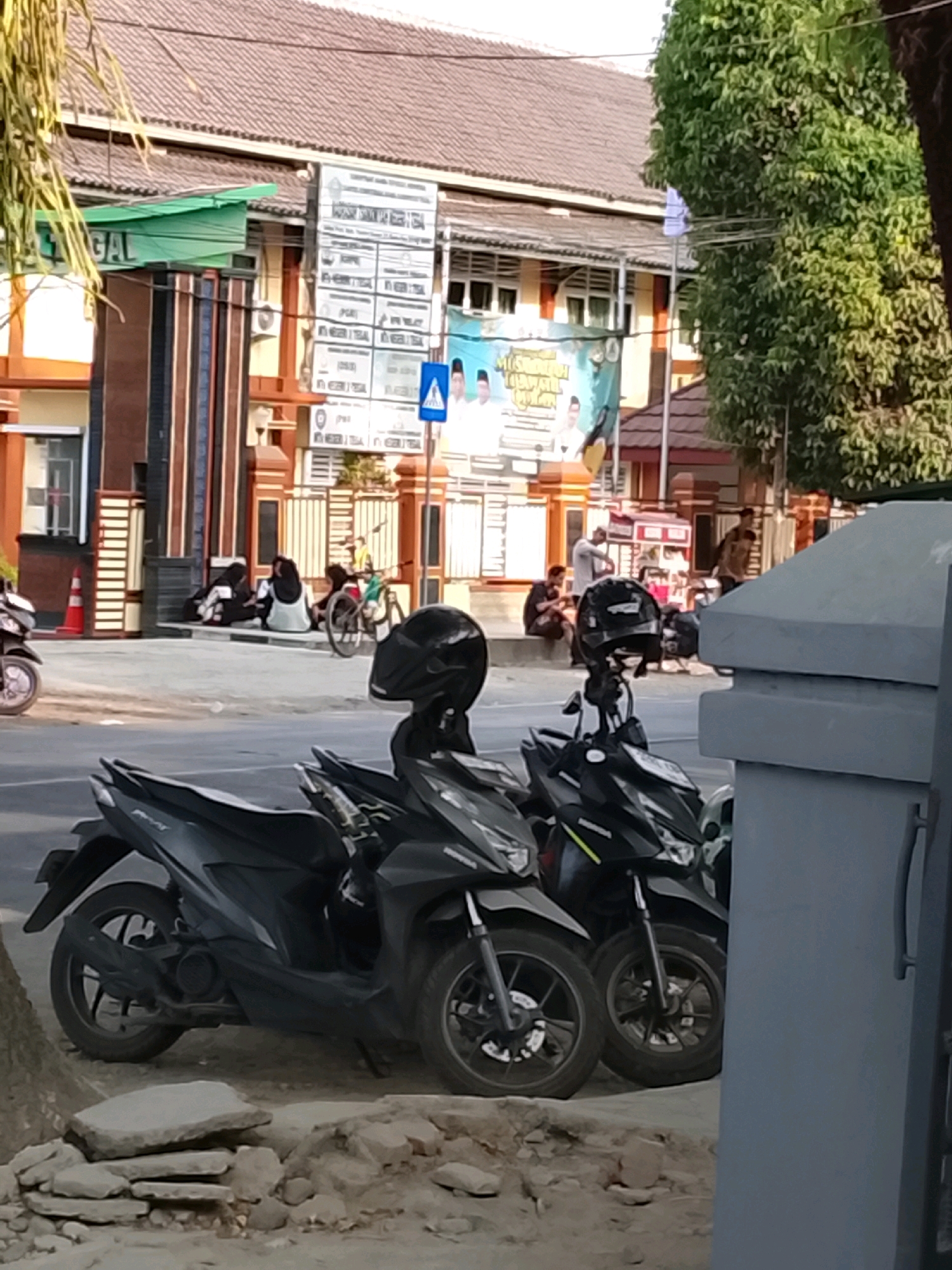 A medium shot captures the exterior of a building, possibly a school or government office, in Slawi, Indonesia. The building features a tiled roof, a green awning, and decorative pillars. In front of the building, several people are seated on a ledge, with a bicycle to their right. A street runs in front of the building, with two black motorcycles parked in the foreground. The motorcycles are facing left and appear to be unattended. The ground in front of the motorcycles is covered in gravel and scattered stones. The scene is well-lit, suggesting it is daytime.